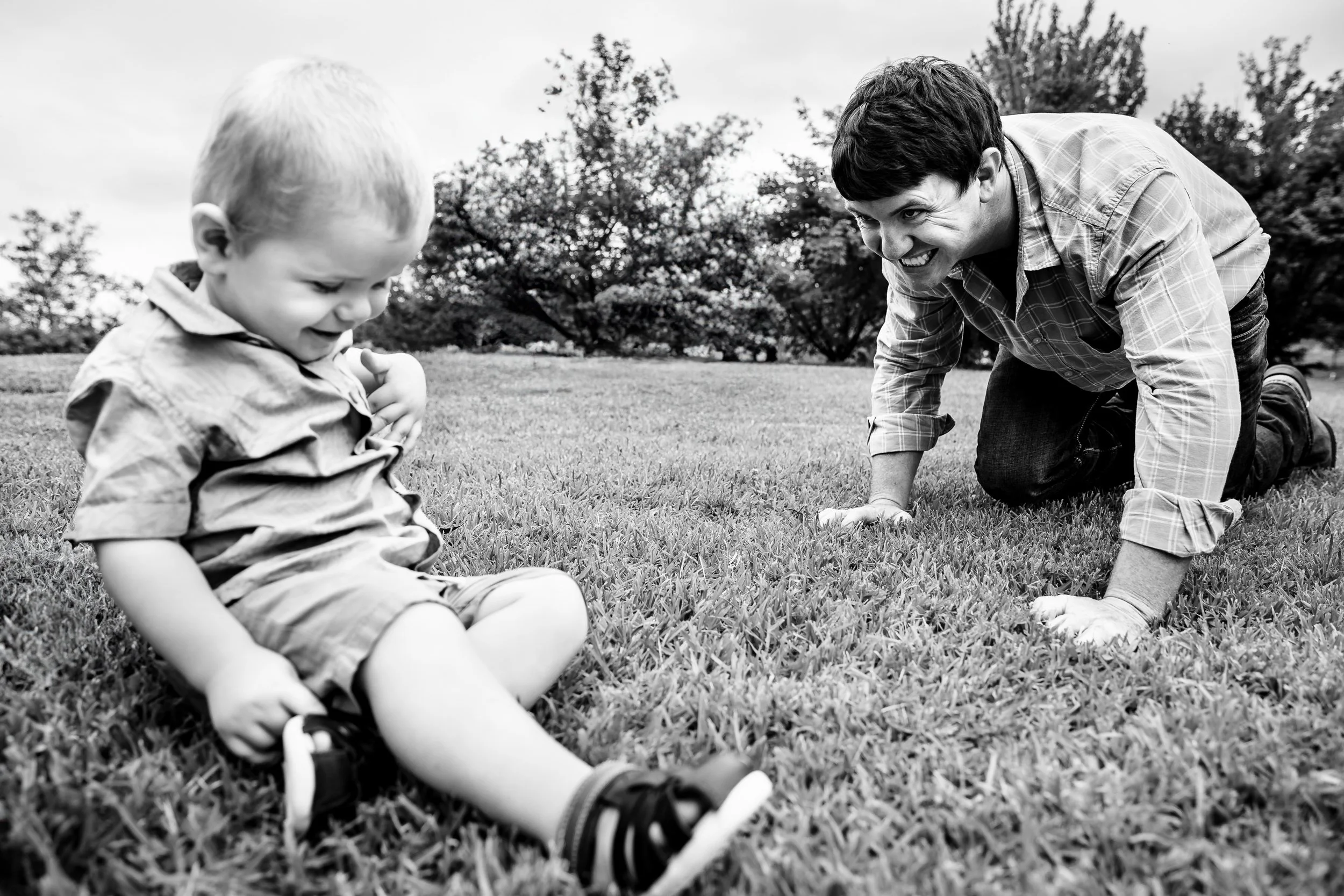 A young man and a small child playing on the grass in a park with trees in the background, smiling and enjoying each other's company.
