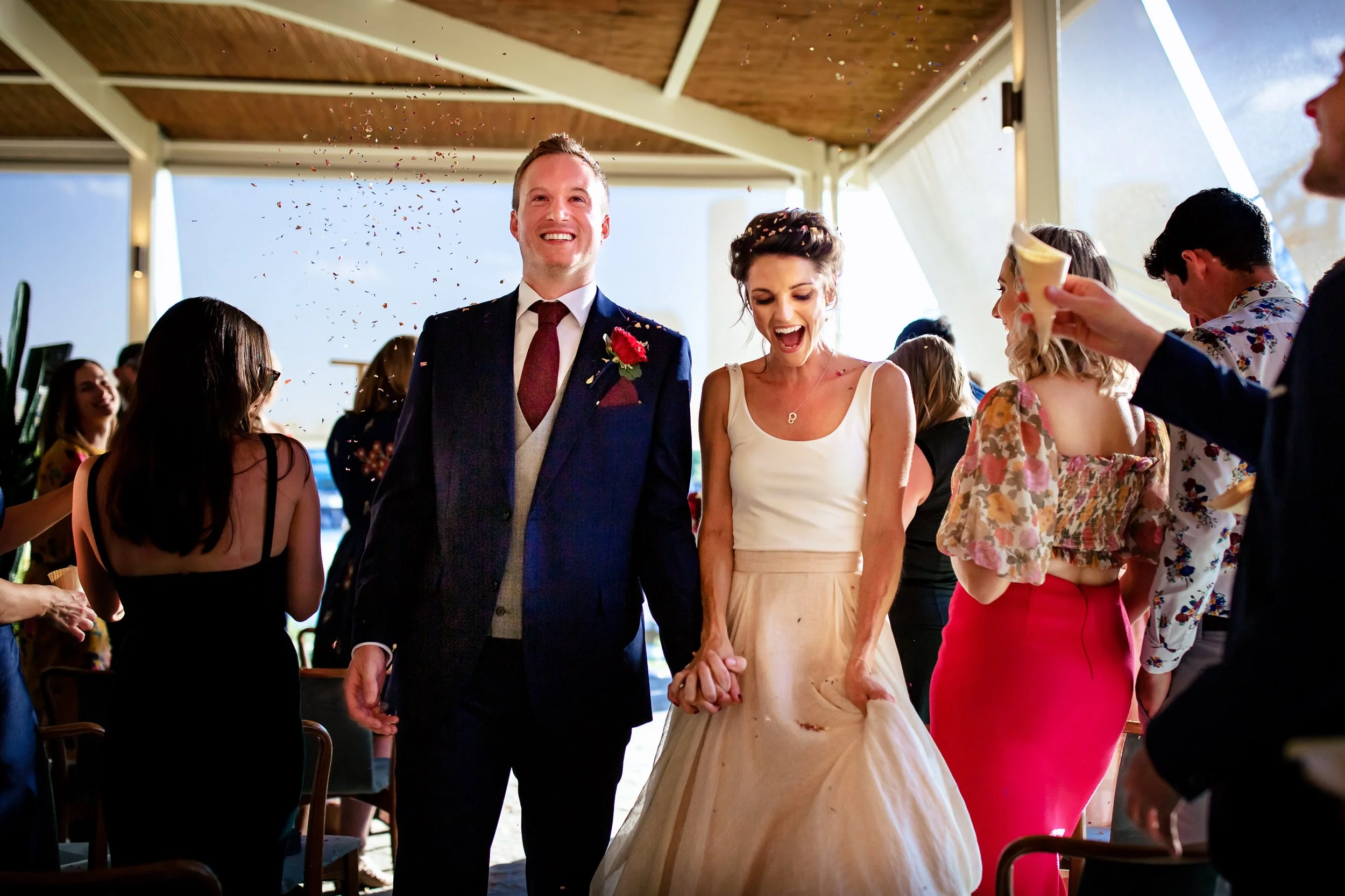 A joyful wedding celebration with the bride and groom holding hands, surrounded by guests under a wooden canopy, some wearing floral attire, and confetti in the air.