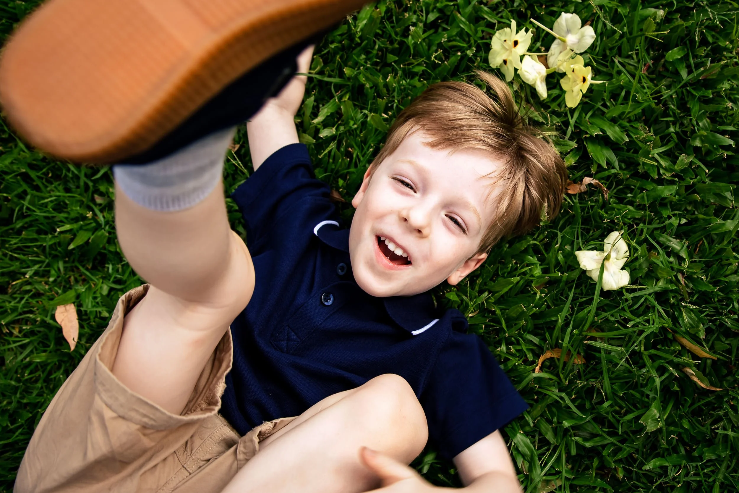 A young boy in Singleton, NSW, lying on grass, smiling, with yellow flowers nearby, holding a basketball.
