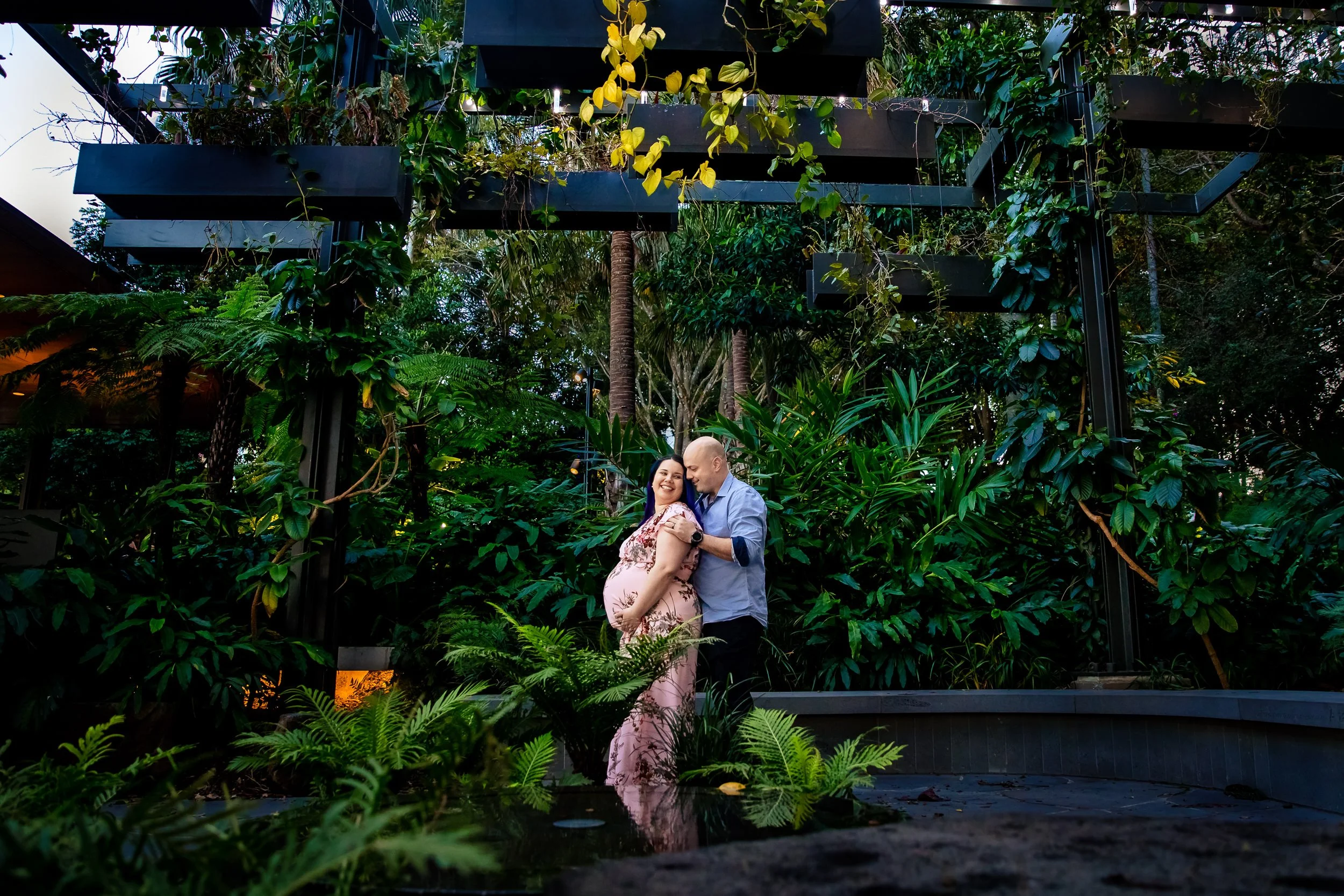 A couple standing close together in a lush, green indoor garden or jungle setting, with the woman pregnant, smiling, and holding her belly, while the man embraces her from the side.