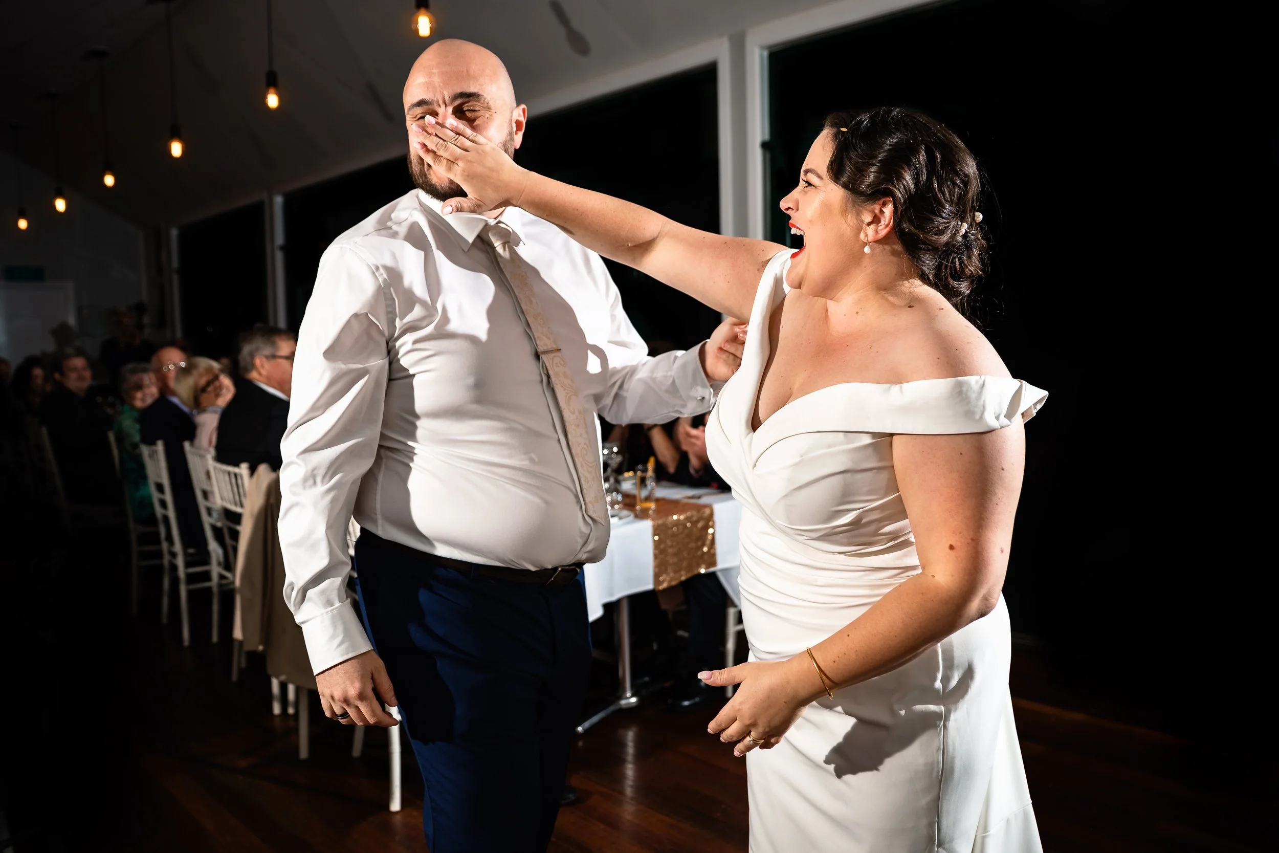 A bride and groom dancing at their wedding reception at Woodlands of Marburg. The bride is shoving cake into the groom's mouth, with guests in the background.