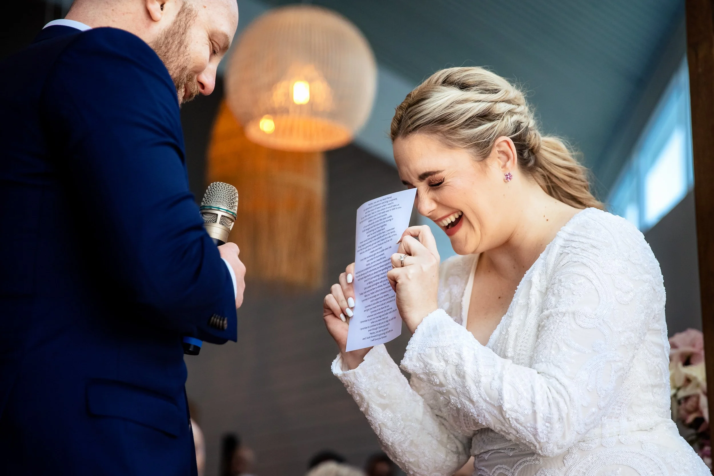 A bride and groom at a wedding ceremony, with the bride laughing as she reads vows from a paper, and the groom holding a microphone, in a softly lit indoor setting. Captured by Lynn & Justin - Hunter Valley & Port Stephens wedding photographers.