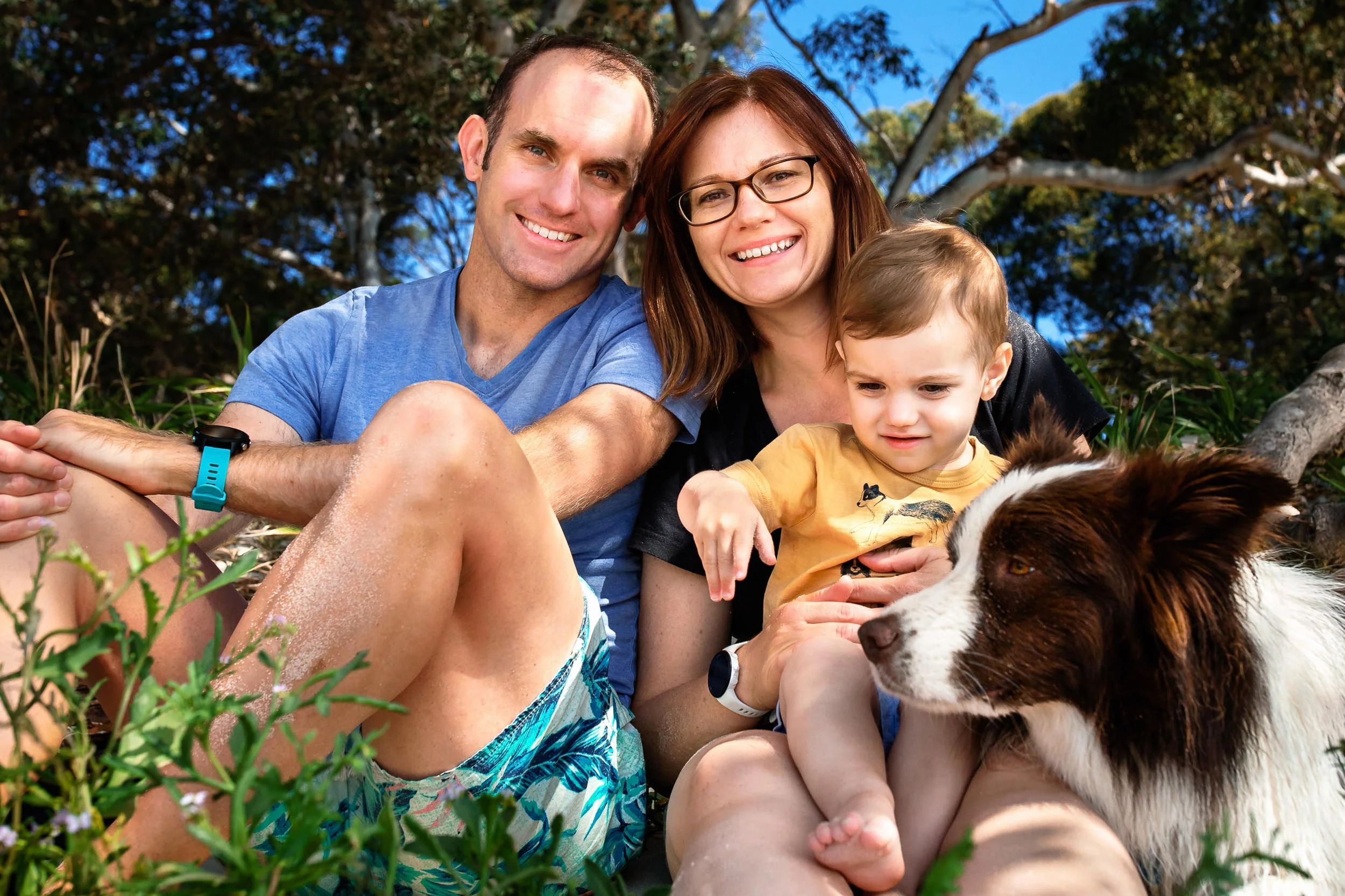 Lynn & Justin - Hunter Valley & Port Stephens wedding photographers - with their family, a dog and a young boy, sitting on Shoal Bay in front of trees, smiling and playing.