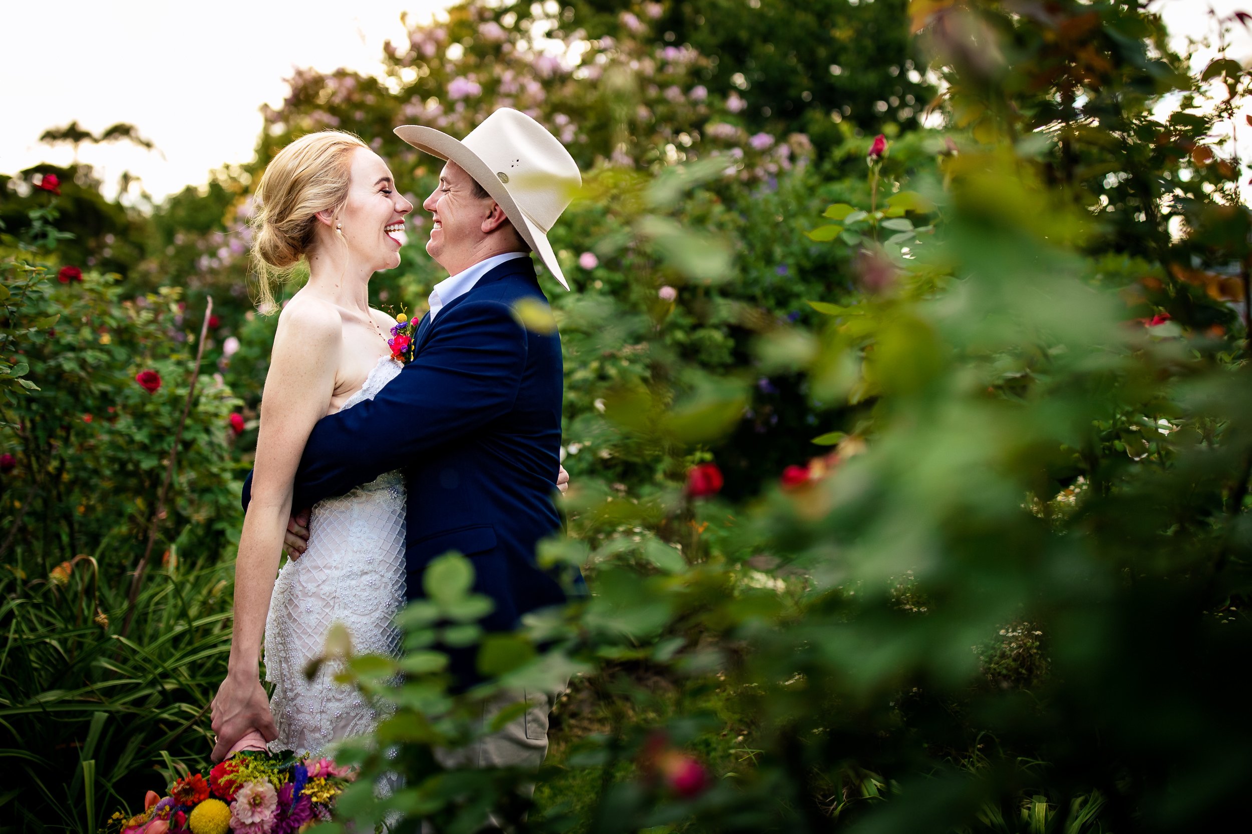 A joyful newlywed couple in Singleton, with the bride in a white lace dress and the groom in a dark suit with a cowboy hat, embracing outdoors among green bushes and pink flowers during sunset, with a bouquet of colorful flowers placed on the ground.