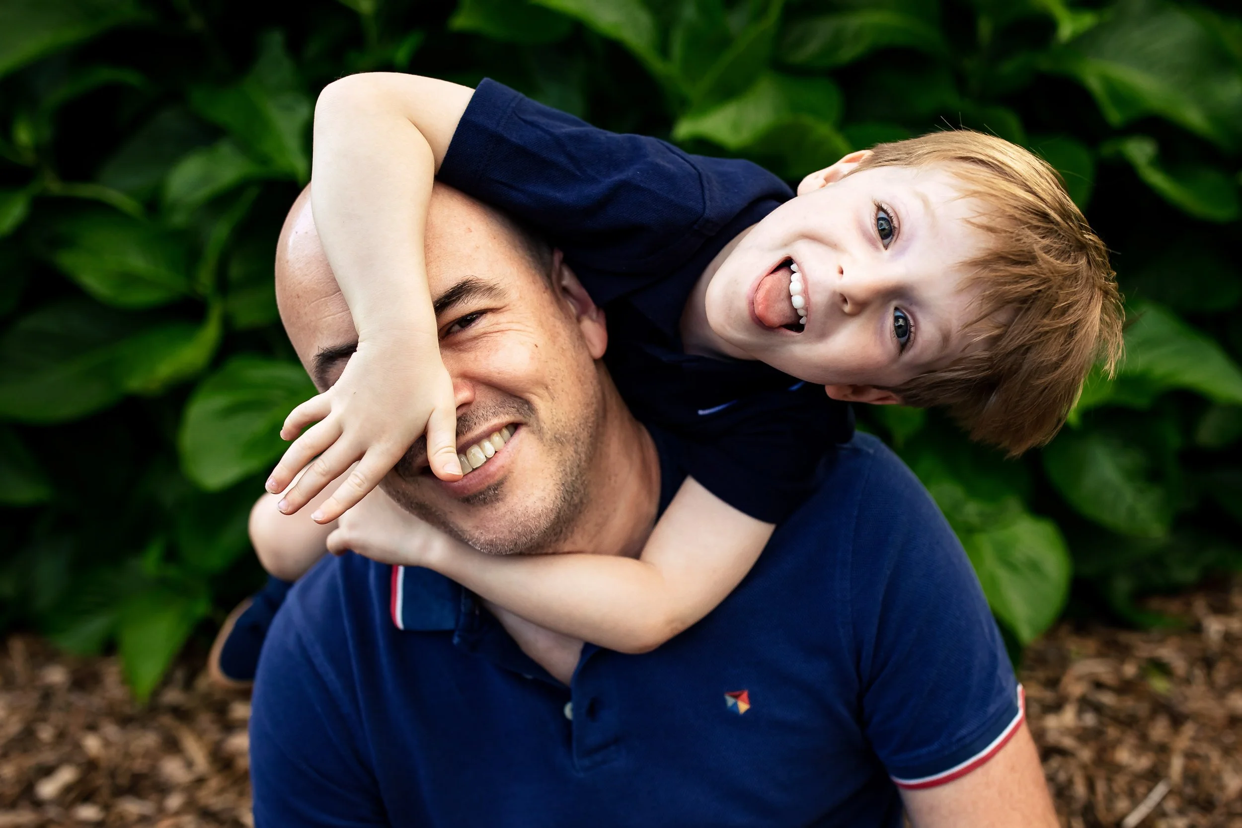A man smiling with a young boy on his shoulders, both outdoors with green foliage in the background.