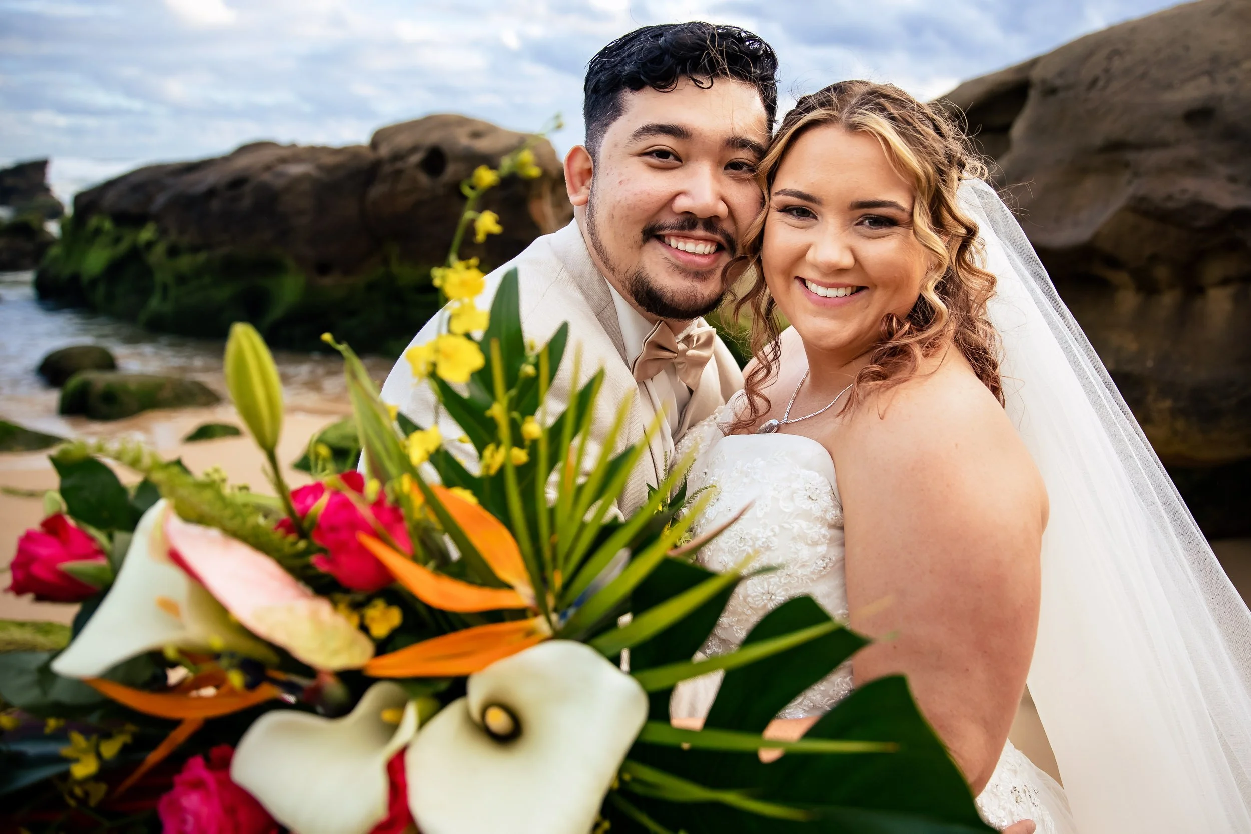 A newlywed couple smiling at the camera, with a Redhead Beach and rocks in the background and a colorful bouquet of flowers in the foreground. Captured by Lynn & Justin, Hunter Valley wedding photographers.