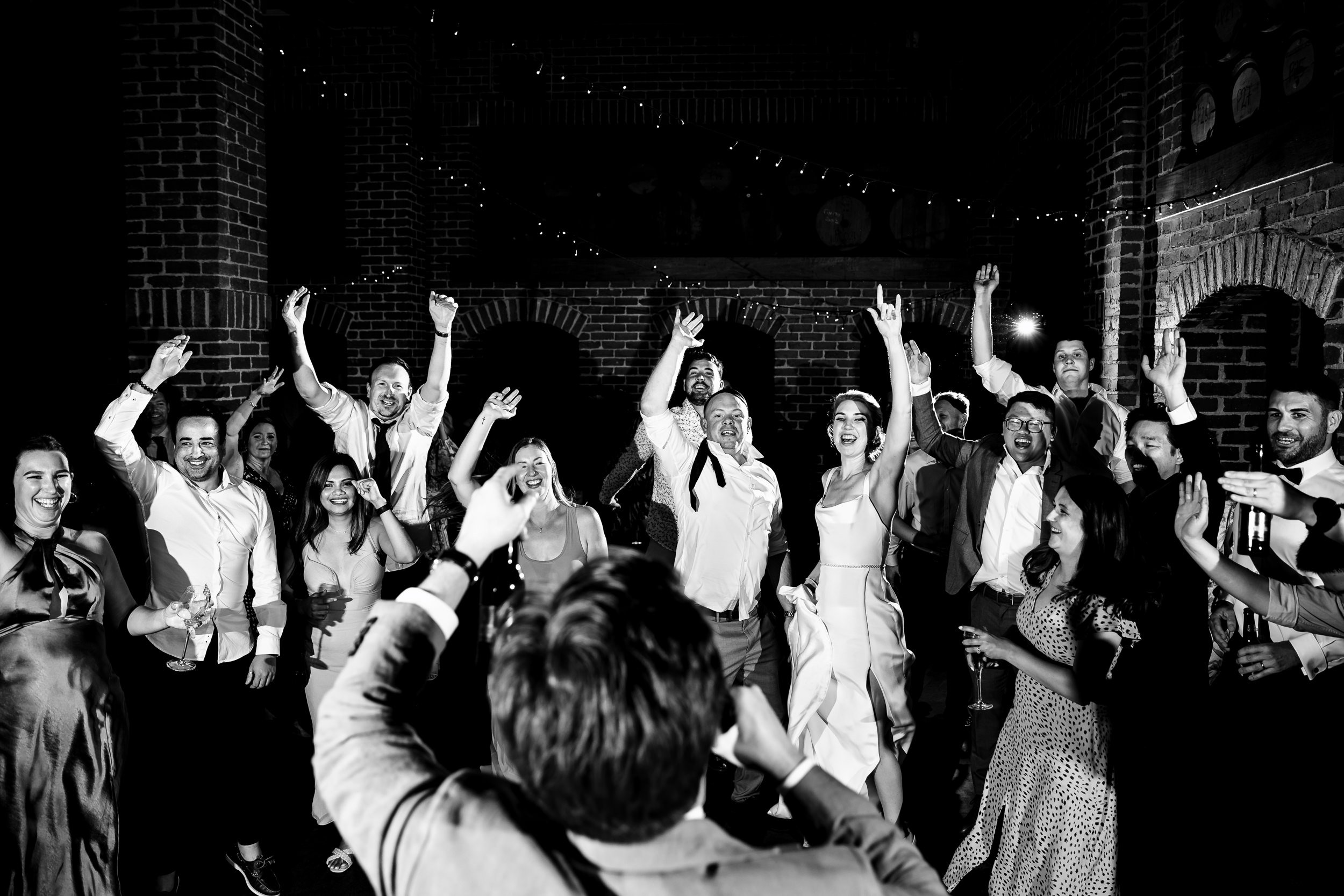 Group of people celebrating at a wedding reception at Sirromet Winery, smiling, dancing, and raising their hands, with a brick wall and string lights in the background. Captured by Port Stephens & Hunter Valley wedding photographers, Lynn & Justin.