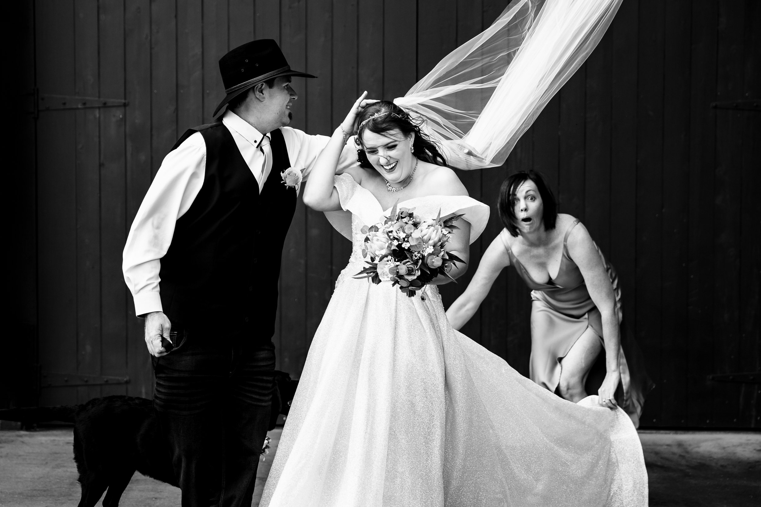 A bride in a wedding dress holding a bouquet, smiling as a man in a suit and hat adjusts her veil, with a woman in a dress to the right reacting with surprise. Captured by Hunter Valley & Port Stephens wedding photographers, Lynn & Justin.
