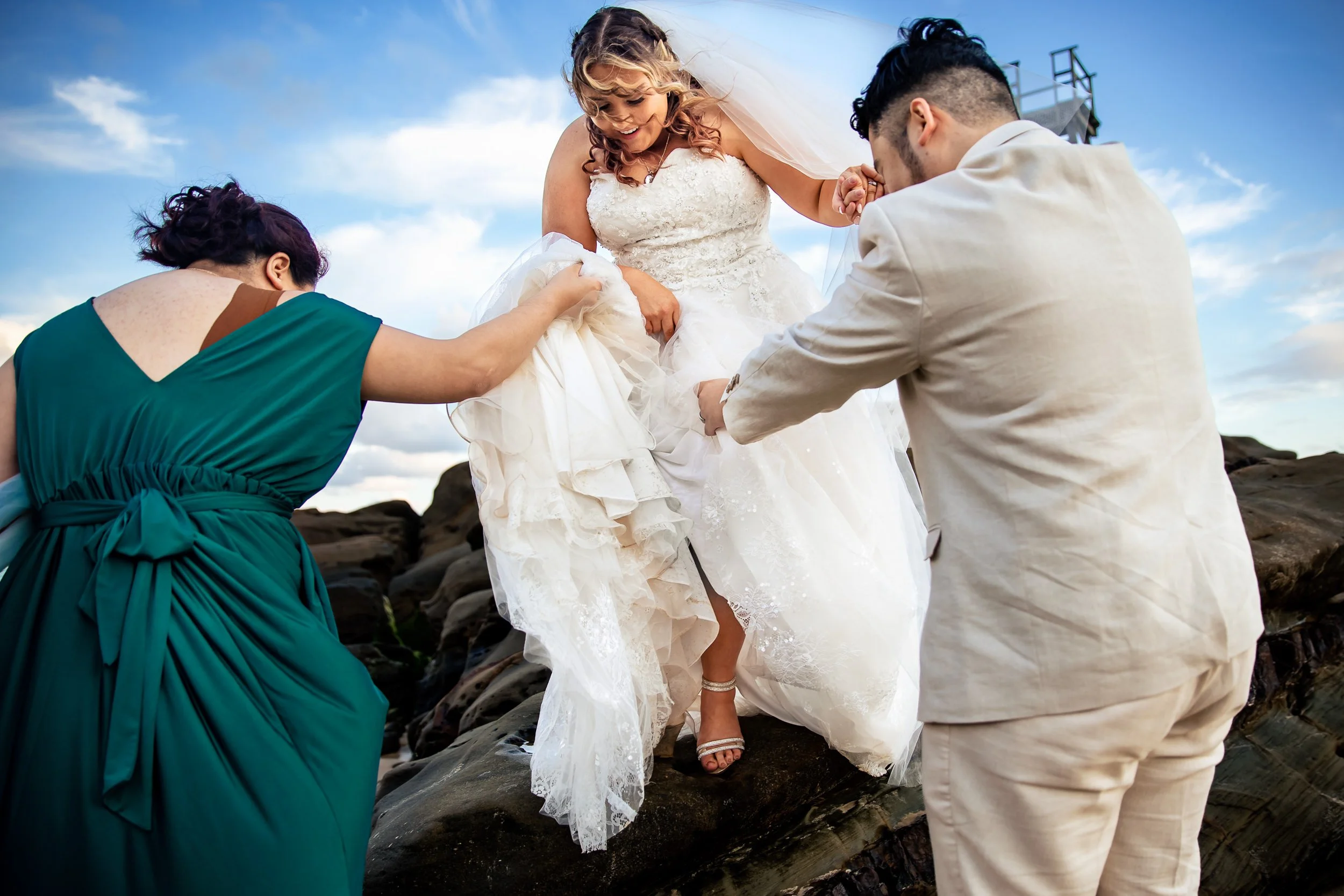 A bride in a white wedding dress stepping over rocks with the help of her groom and bridesmaid during at Redhead Beach, Newcastle. Captured by Hunter Valley wedding photographers, Lynn & Justin.