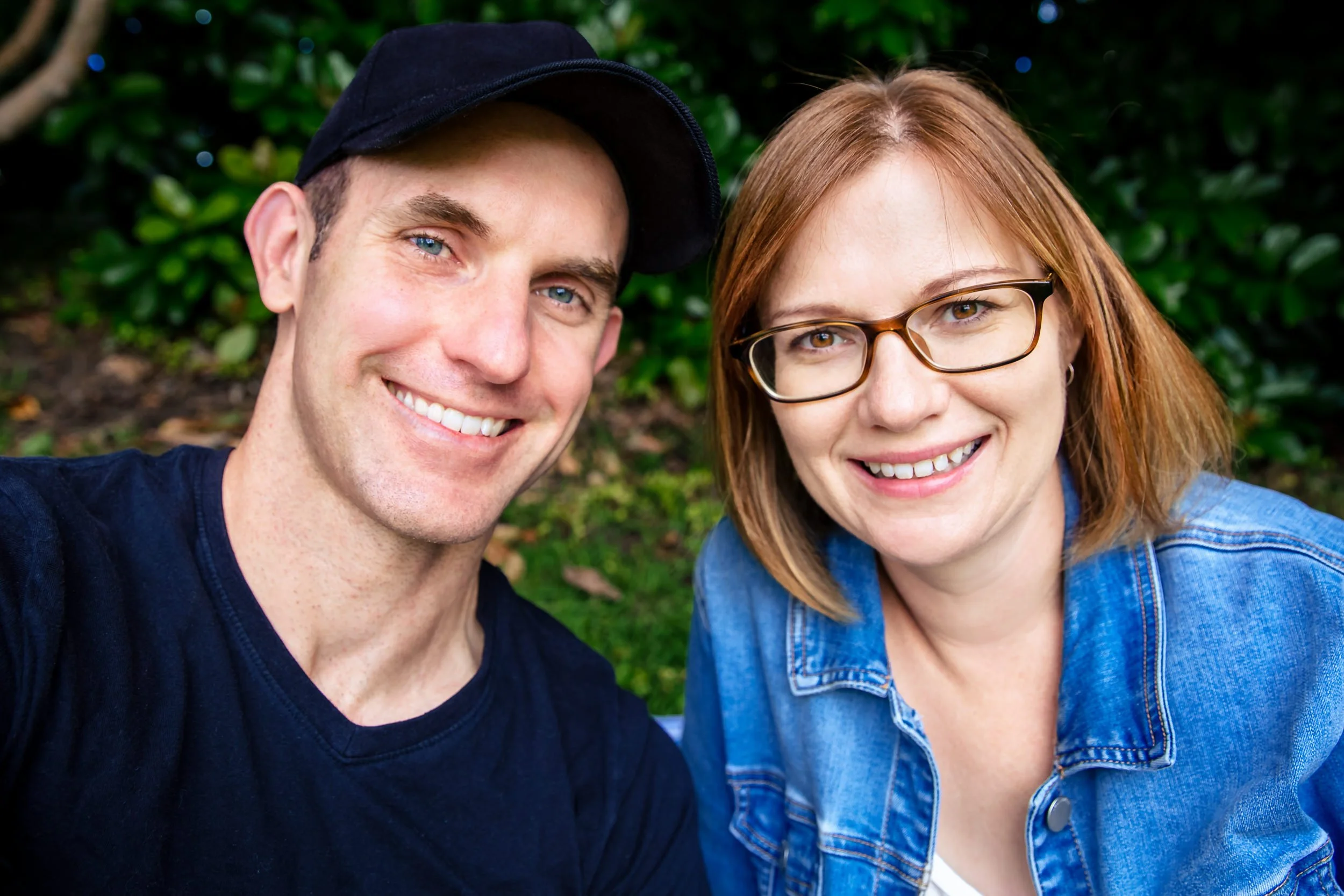 A young man with blue eyes wearing a black cap and a young woman with glasses and red hair wearing a denim jacket, both smiling and taking a selfie outdoors with green foliage in the background.  Taken in Singleton, NSW.