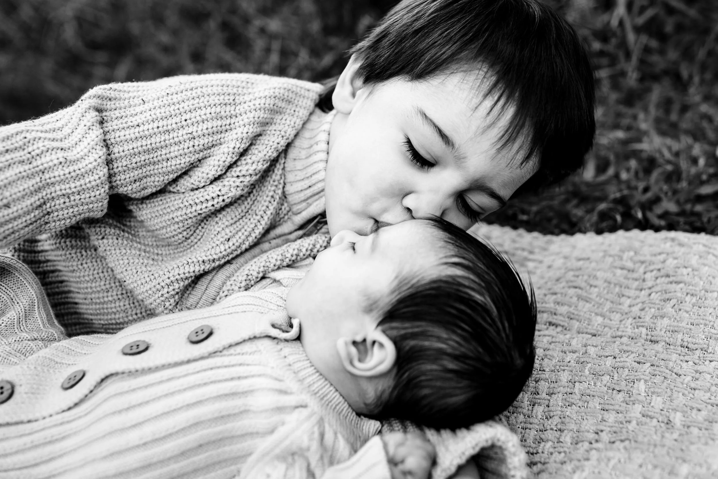 A young boy with short dark hair lying on the ground, gently touching foreheads with a small girl with long dark hair, their eyes closed, in a moment of tender connection.