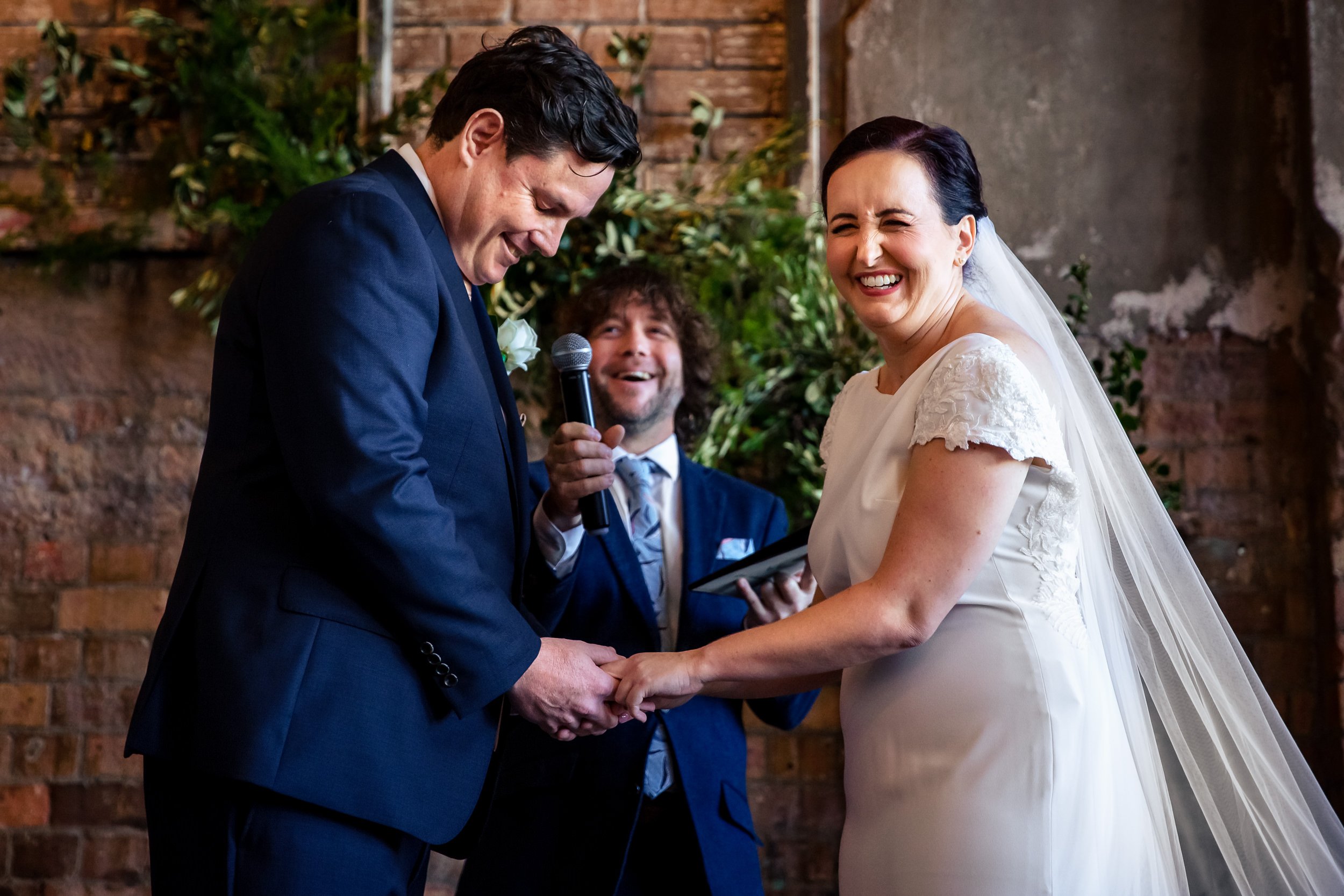 A bride and groom holding hands and exchanging vows during their wedding ceremony at The Warehouse, Brisbane, smiling and laughing, with a officiant in the background.