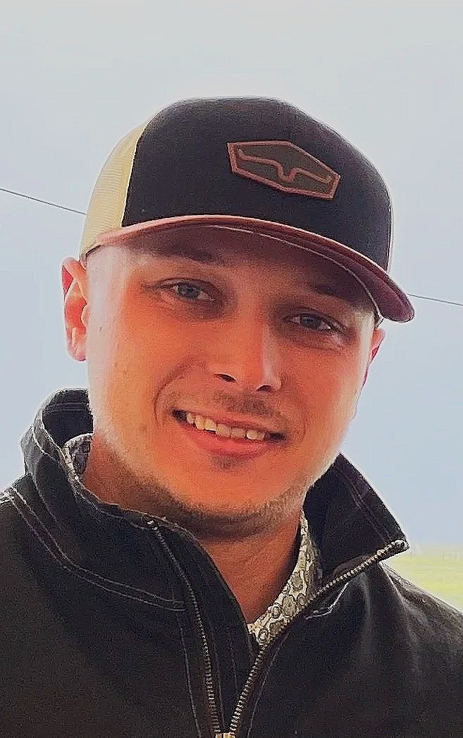 Young man wearing a black and beige trucker hat and black jacket, smiling outdoors.