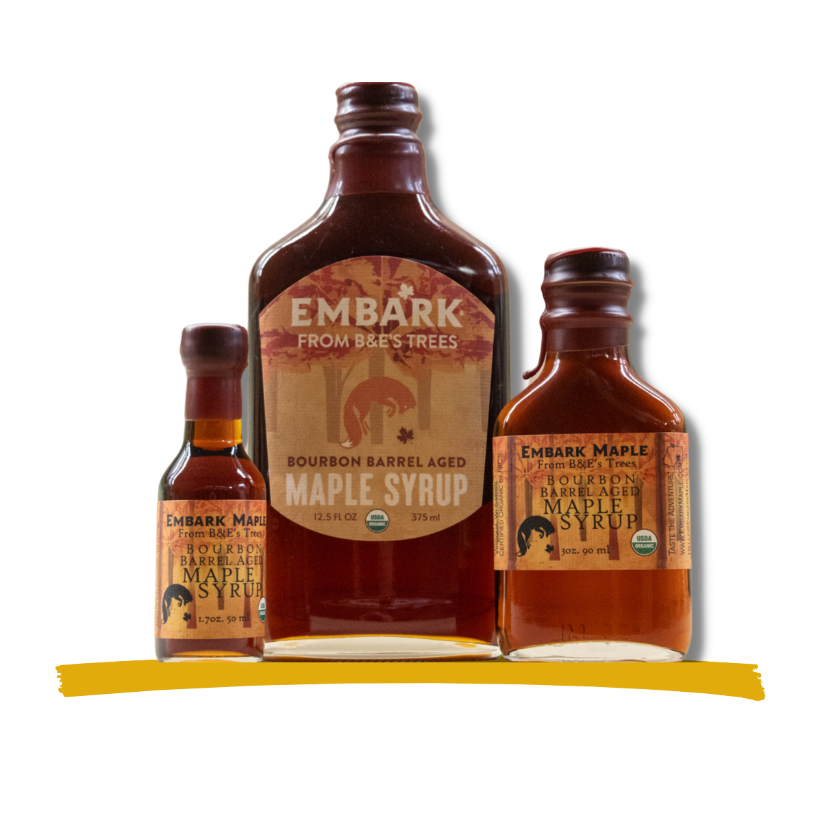 Three bottles of Embar Maple Syrup, varying in size, with labels indicating bourbon barrel aged maple syrup from B&E's Trees. The labels are orange with black and white text. The largest bottle is in the center, flanked by two smaller bottles on either side, all placed on a yellow surface against a black background.