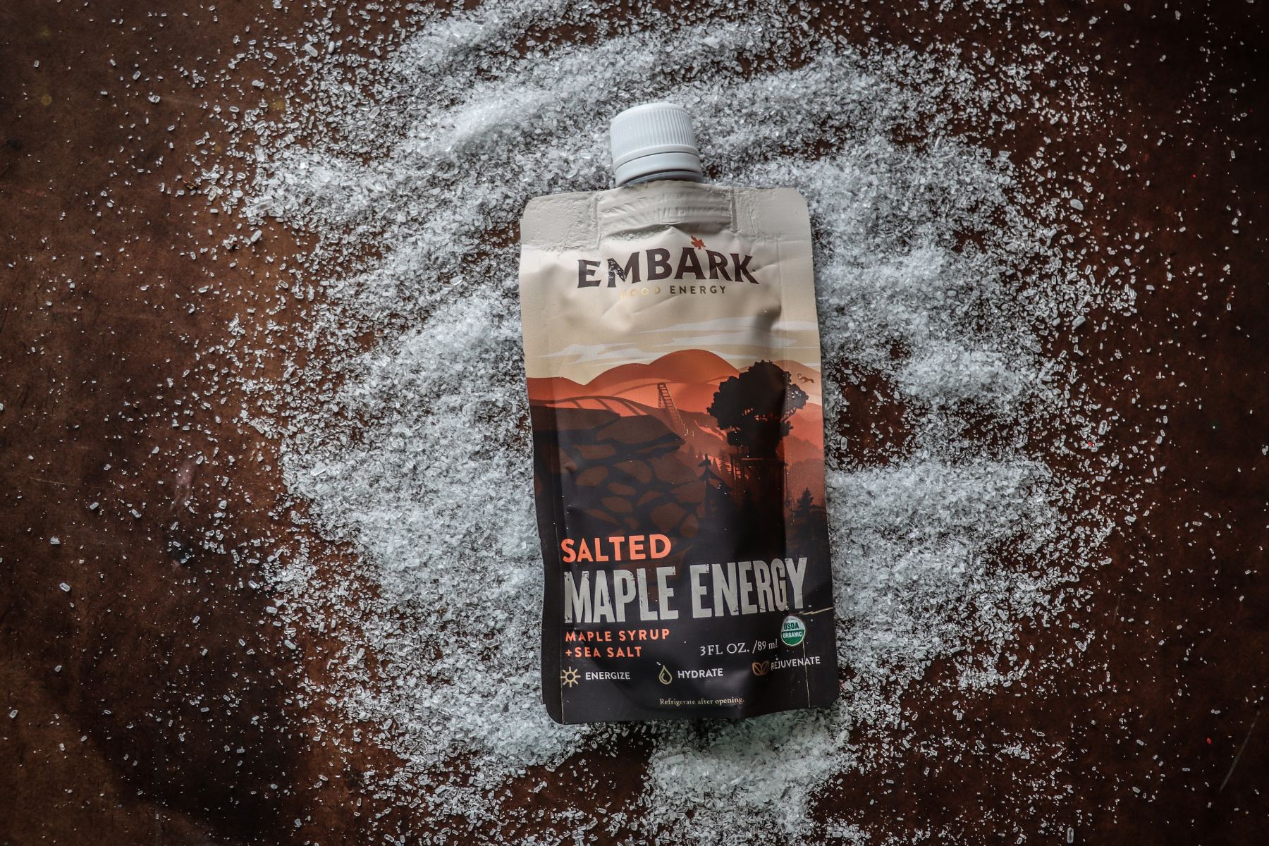 A beverage pouch labeled 'Embark Salted Maple Energy' placed on a dark surface surrounded by scattered sea salt.