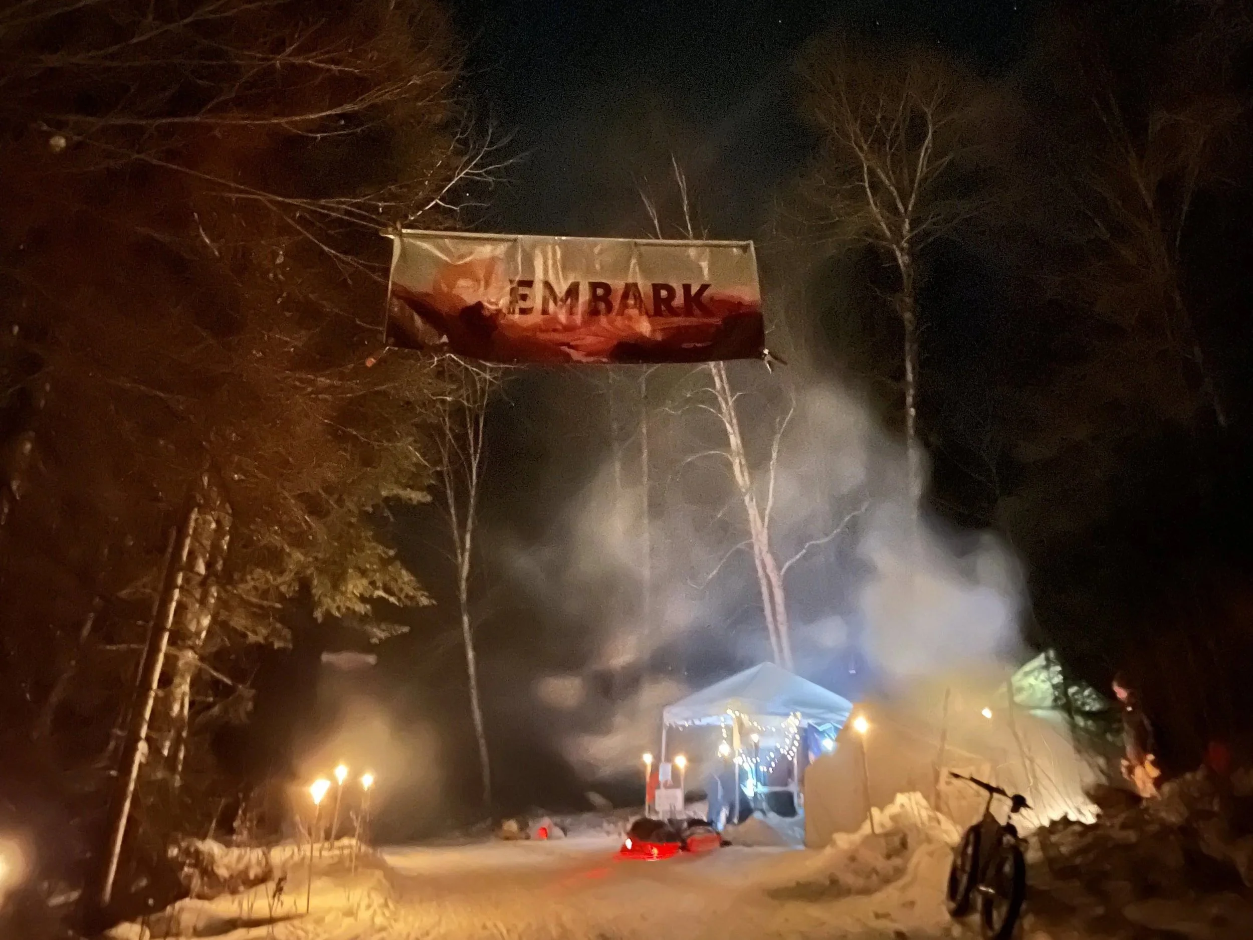 Night scene of a street with trees and vehicles, illuminated by streetlights, with a large banner that reads 'EMBARK' hanging between the trees.