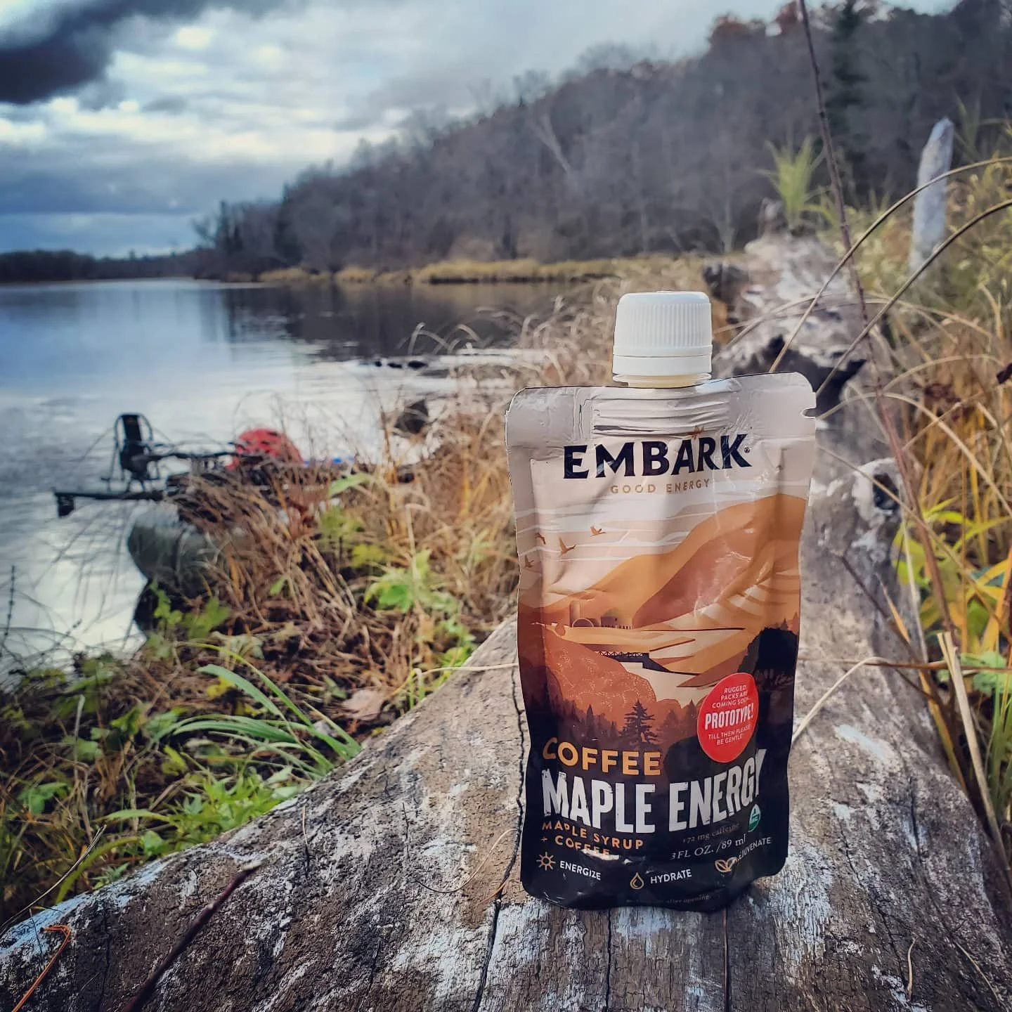 A pouch of Embark Coffee Maple Energy on a log near a riverbank with packrafts and wooded hills in the background.