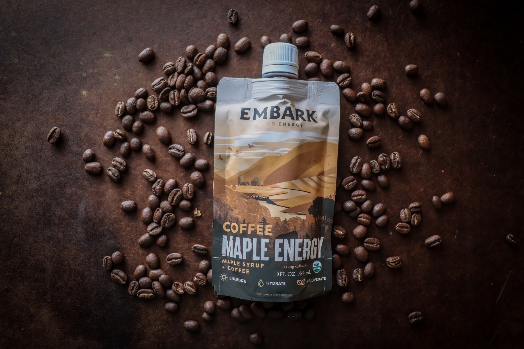 A pouch of Embark Coffee Maple Energy drink surrounded by scattered coffee beans on a dark surface.