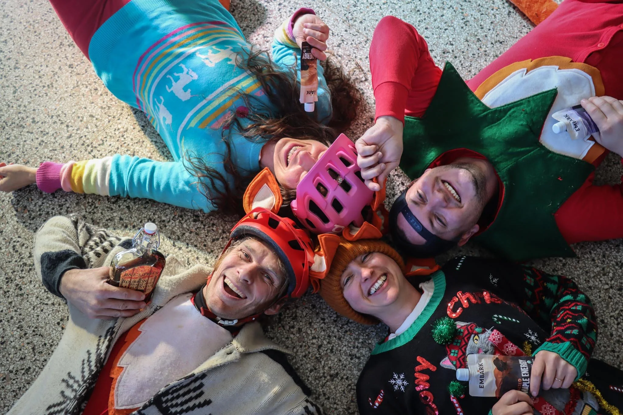 A group of people lying on the floor celebrating Christmas. They are wearing festive holiday sweaters and hats, some holding bottles of syrup. They are smiling and appear to be happy.