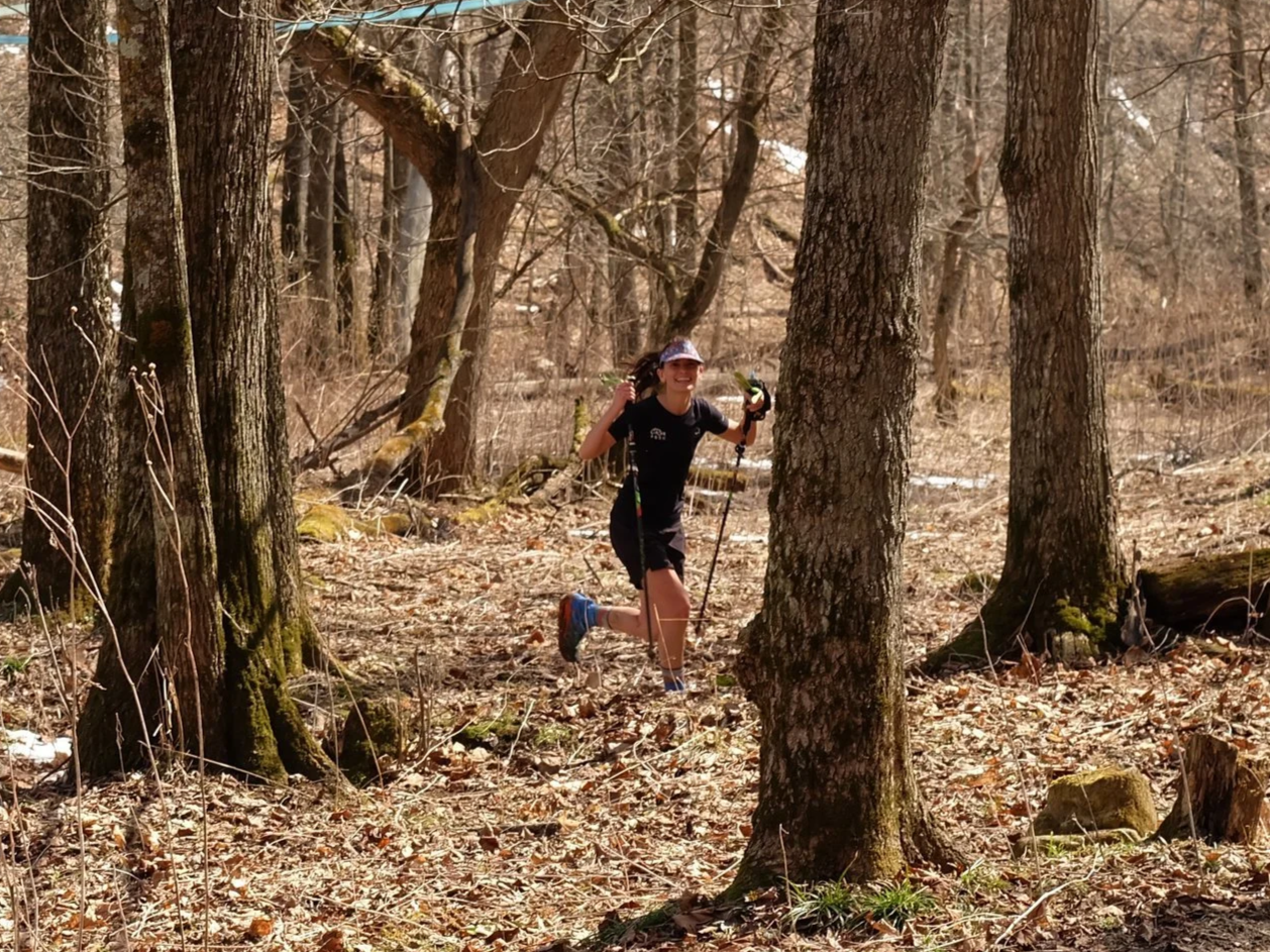 A woman trail running in a wooded forest during daytime, wearing black athletic clothing, hat, and sunglasses, holding trekking poles.