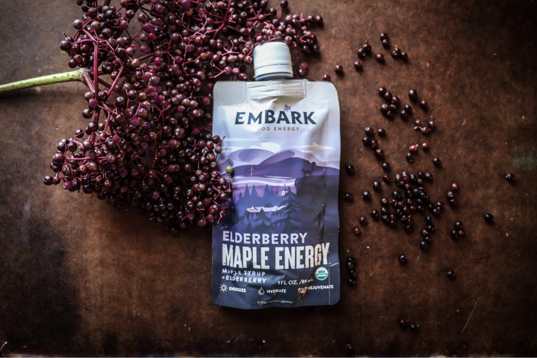 A purple Elderberry Maple Energy drink pouch placed on a brown surface, surrounded by fresh elderberry clusters and scattered elderberries.