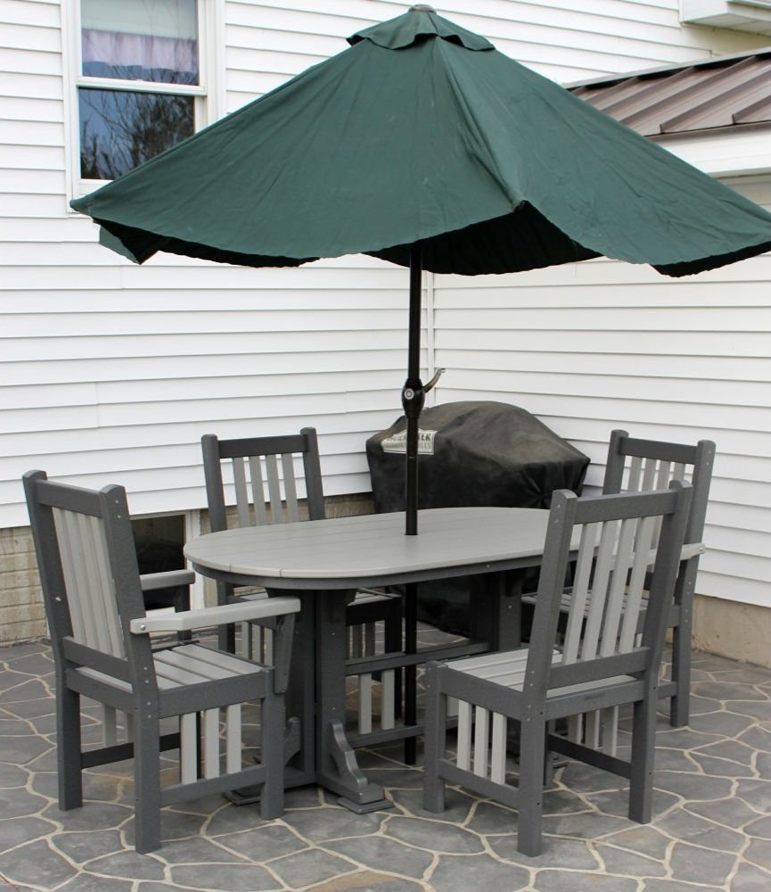 Outdoor patio set with a table, four chairs, and a green umbrella, placed on a tiled surface next to a white house and a covered grill.