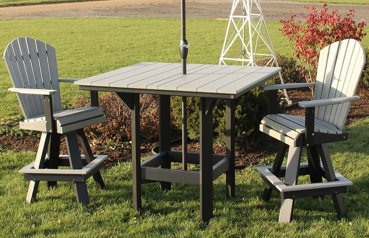 Outdoor patio set with two gray Adirondack chairs and a square table on grass, surrounded by greenery and a white windmill structure in the background.