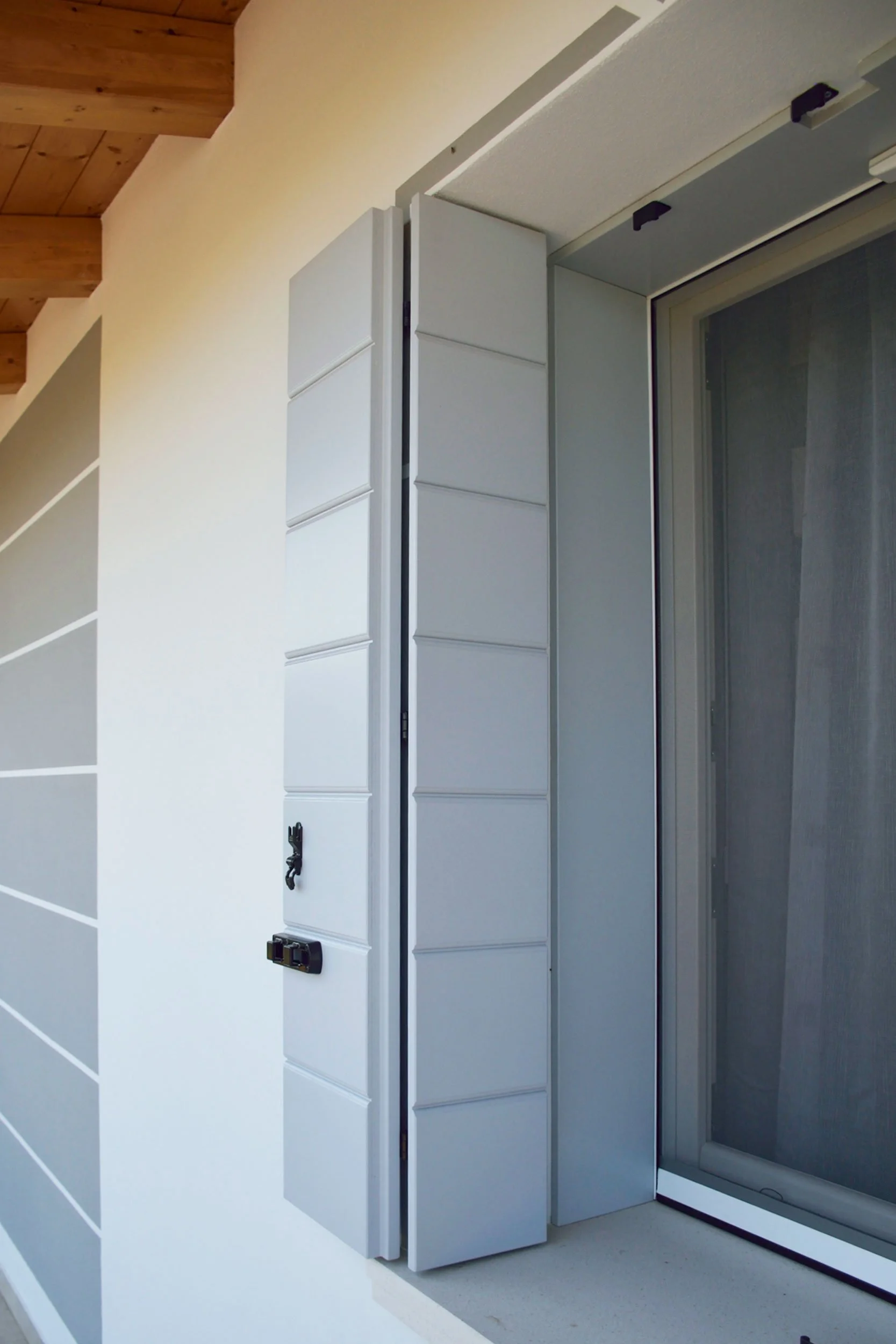 Particolare di una finestra con tapparelle in posizione abbassata e un pannello di rivestimento bianco vicino, sotto un soffitto in legno.