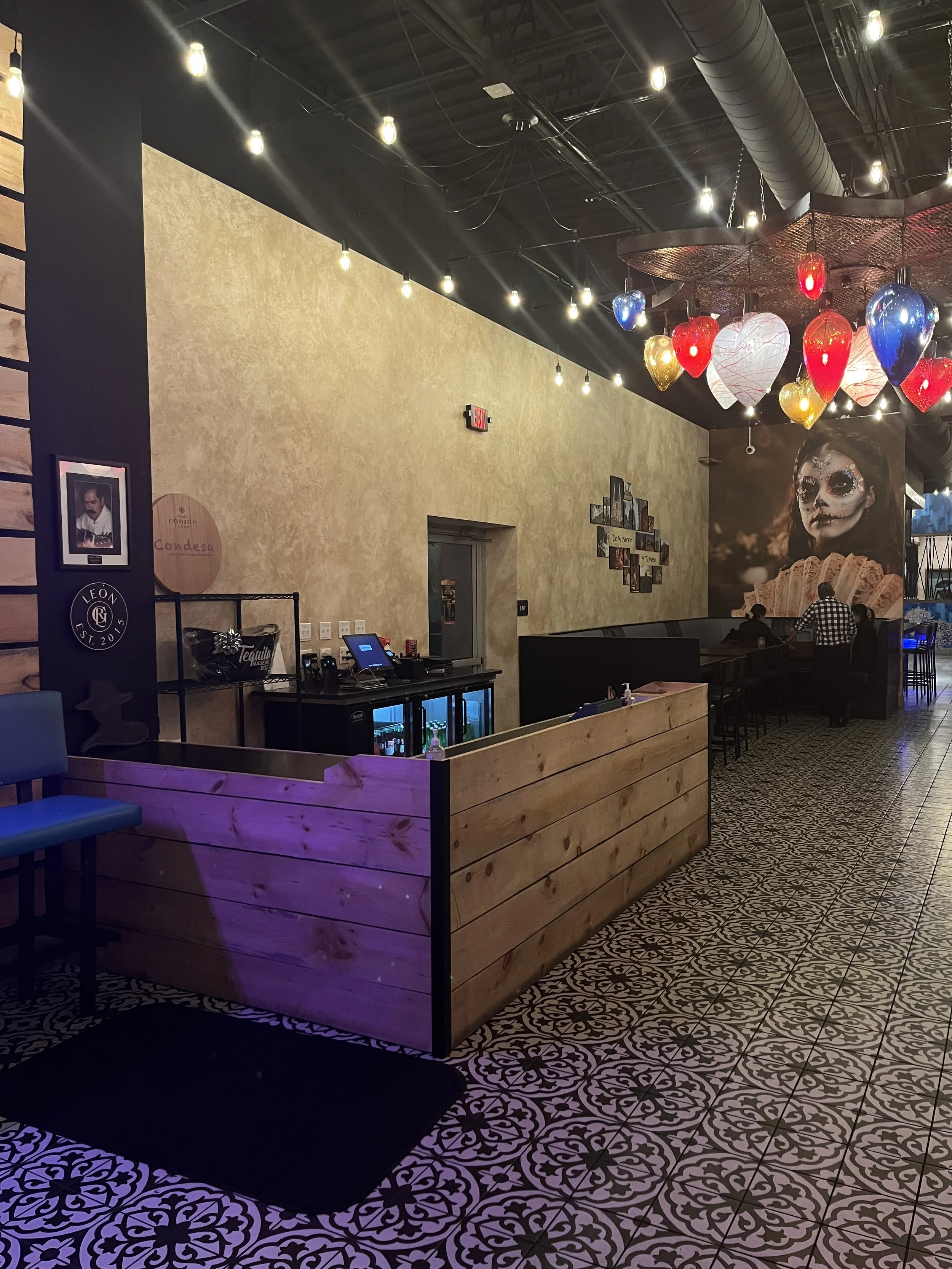 Interior of a restaurant with patterned tile flooring, wooden reception counter, colorful hanging lights, and a mural of a woman with face paint.Home show kitchen floors checkerboard faux painted floor venetian plaster metallic designer finishes pain