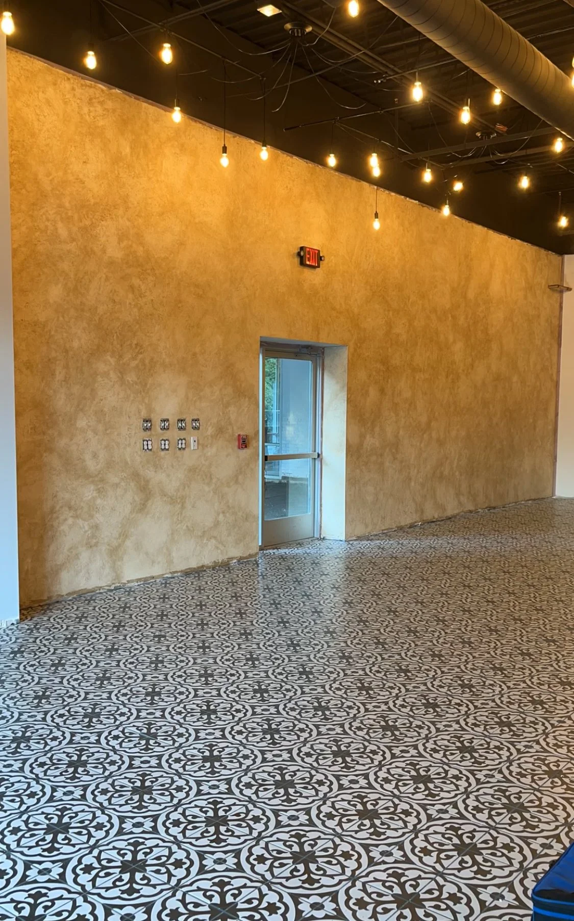 Interior of a building with ornate tile flooring, textured beige wall, hanging light bulbs, and a glass exit door.Home show kitchen floors checkerboard faux painted floor venetian plaster metallic designer finishes paint glazes 