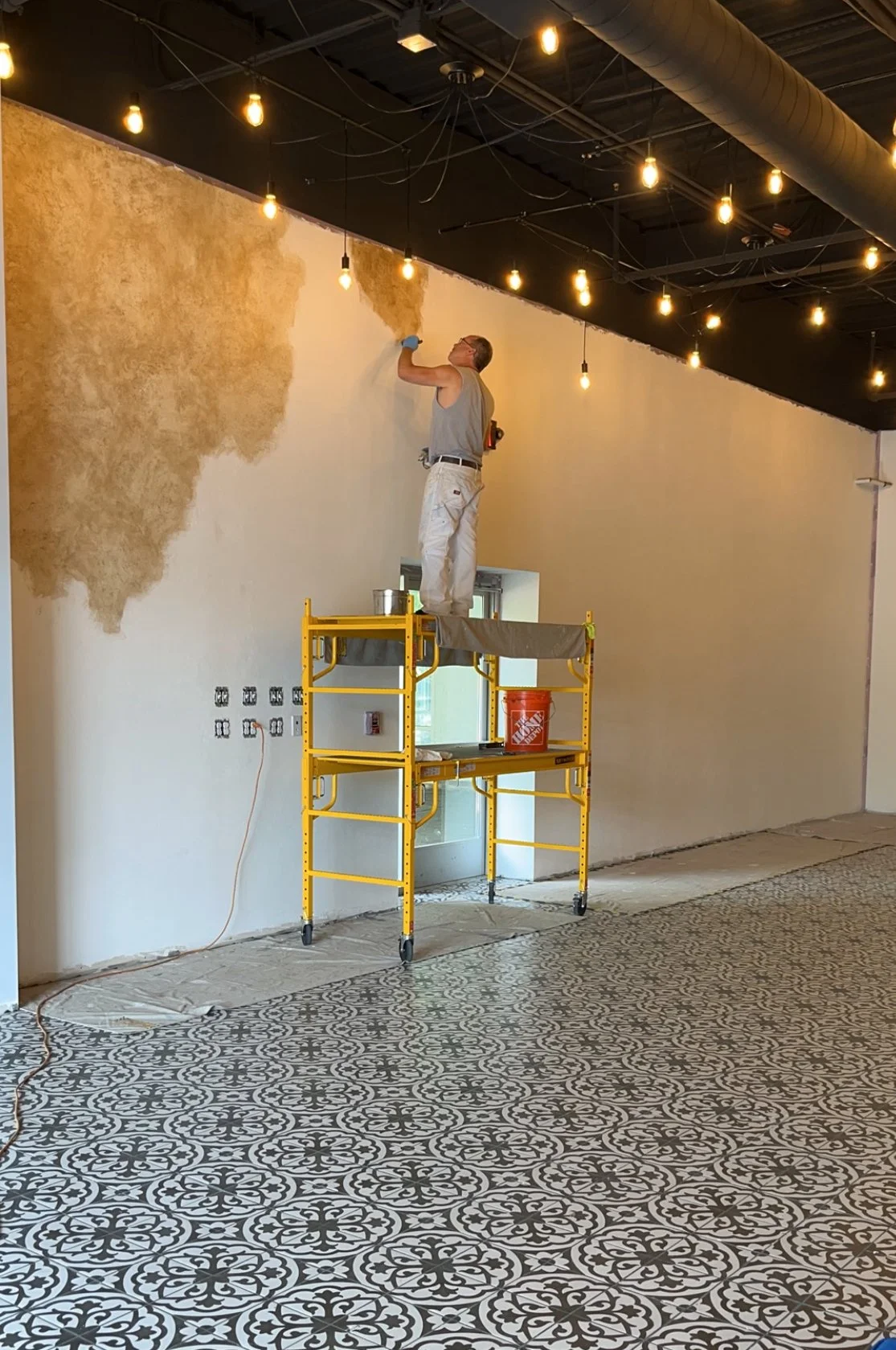 Interior painting in progress; person on scaffolding painting wall with brown texture, hanging lights, patterned tile floor.Home show kitchen floors checkerboard faux painted floor venetian plaster metallic designer finishes paint glazes 