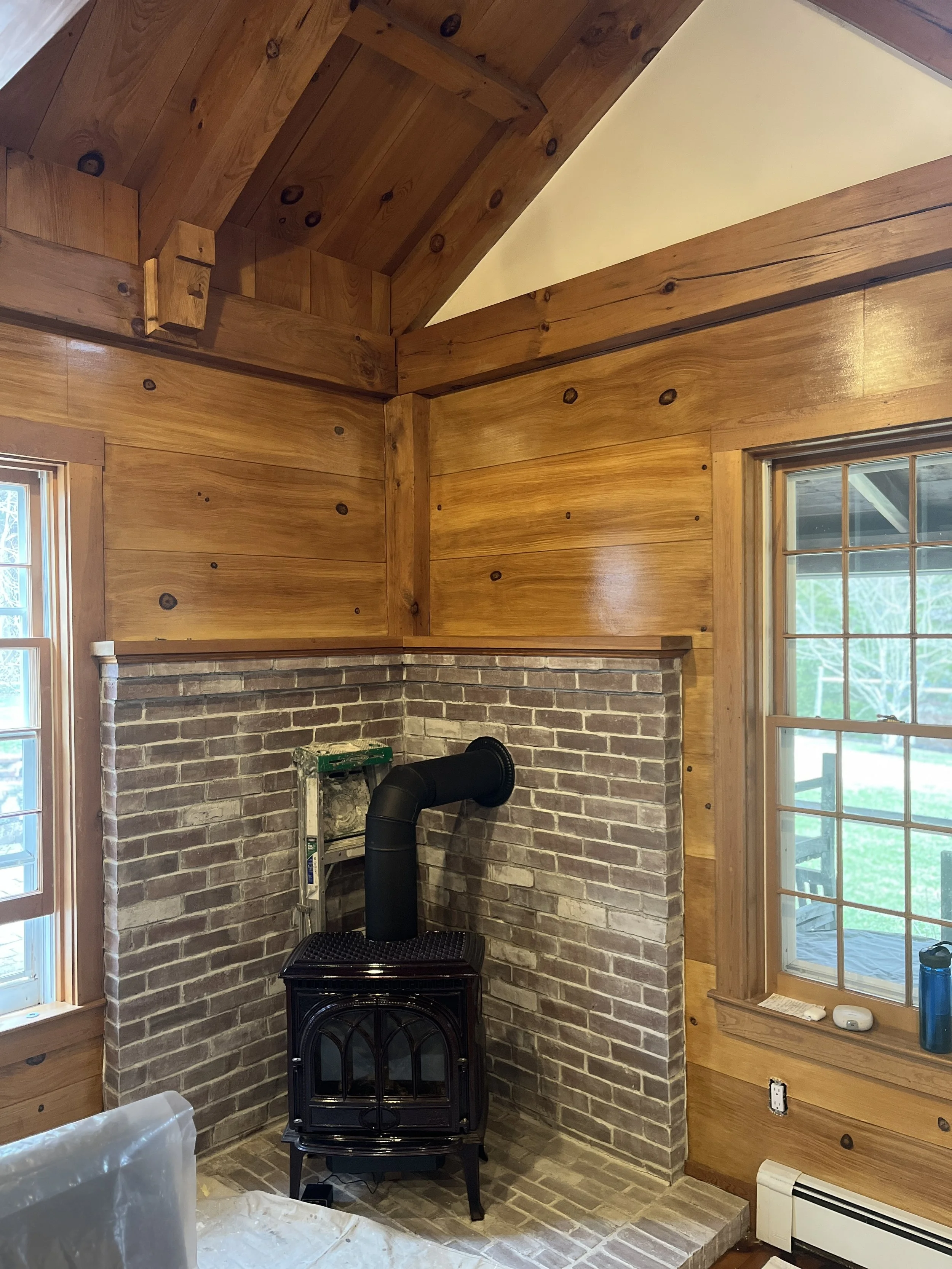 Wood-paneled room with a corner featuring a black wood-burning stove and brick wall surround. Large windows on both sides.Home show kitchen floors checkerboard faux painted floor venetian plaster metallic designer finishes paint glazes 