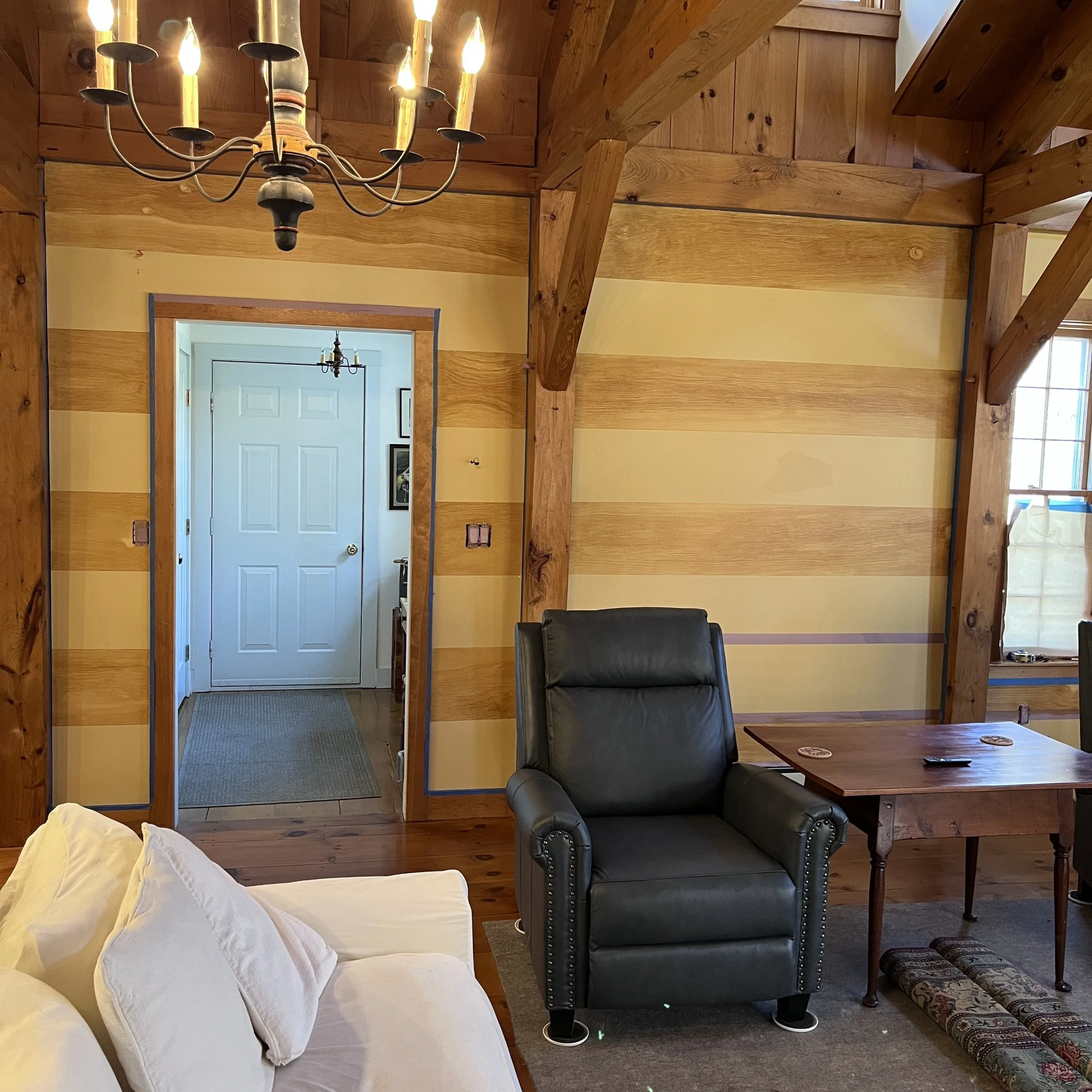 Cozy cabin-style living room with wooden beams, a white sofa, and a black leather recliner. A wooden table is to the right, with a simple chandelier hanging from the ceiling. A doorway leads to a different room with a white door and framed art.Home s