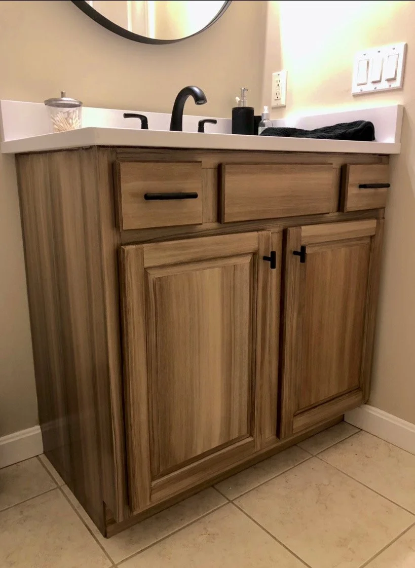 Bathroom vanity with wooden cabinets, black faucet, round mirror, soap dispenser, and small canister on tiled floor.Home show kitchen floors checkerboard faux painted floor venetian plaster metallic designer finishes paint glazes 
