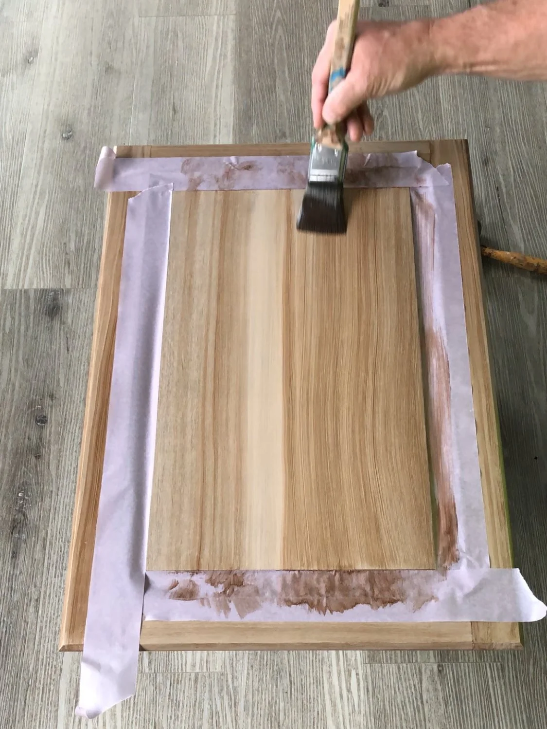 Person applying stain to wooden panel using a brush, with painter's tape along the edges.Home show kitchen floors checkerboard faux painted floor venetian plaster metallic designer finishes paint glazes 