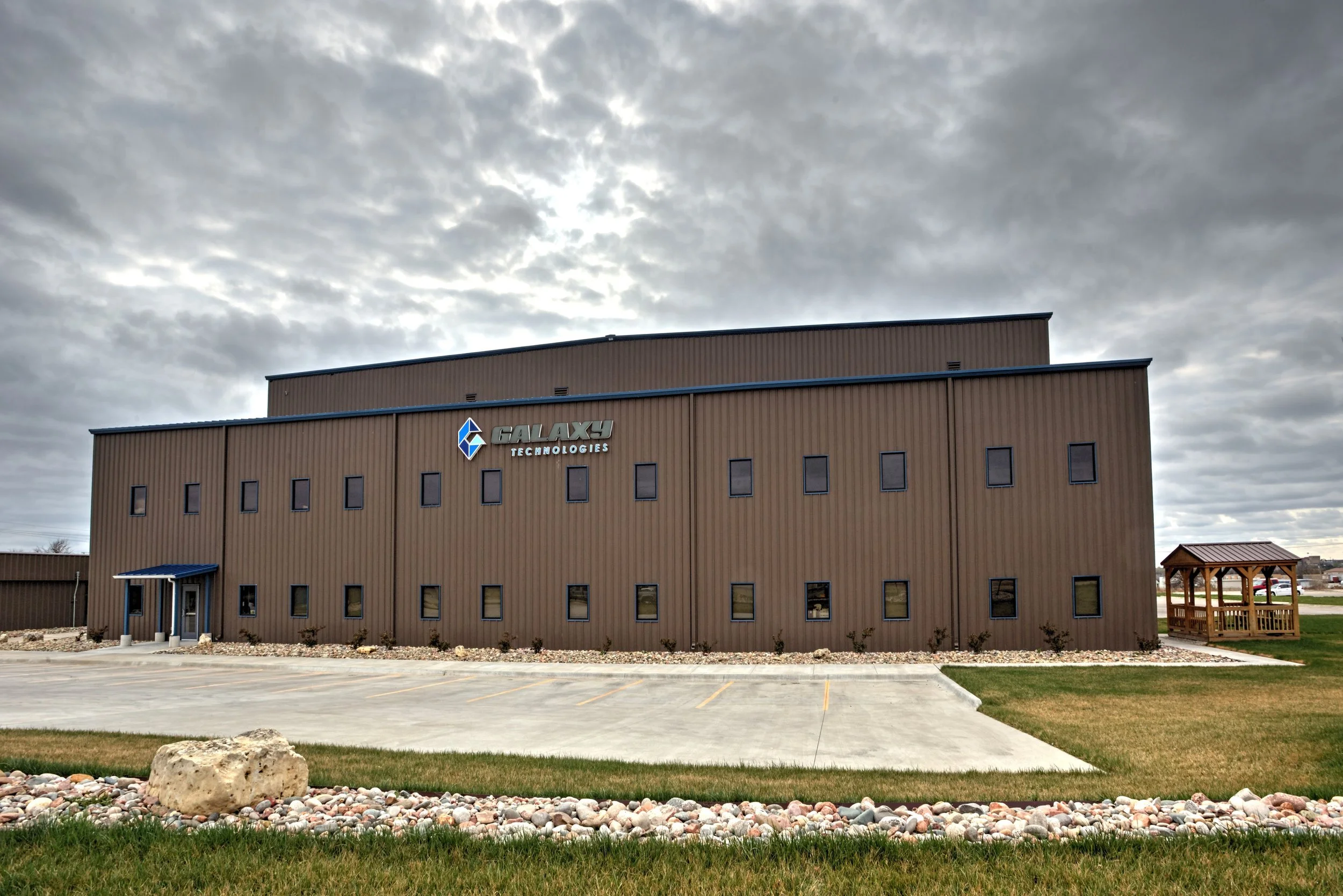 Exterior of a modern, brown industrial building with a blue roof overhang, displaying the 'Galaxy Technologies' sign. The building has many small windows and is surrounded by a parking lot, a small grassy area, and a wooden gazebo on the right side with cloudy skies overhead.