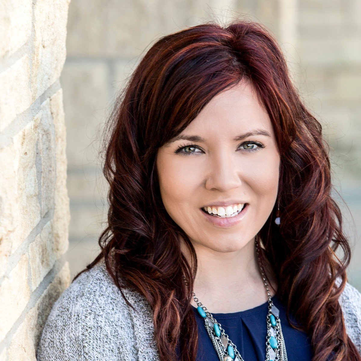A woman with red hair and blue eyes smiling, wearing a necklace and a gray sweater, standing outdoors near a brick wall.