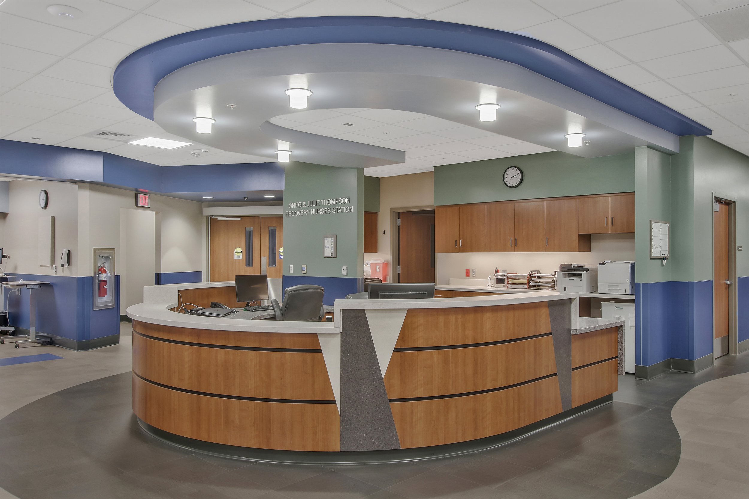 A hospital reception desk with a curved wooden counter, a computer, and chairs. The background includes a sign that reads "GREG & JULIE THOMPSON RECOVERY NURSES STATION," a clock, cabinets, and doors, with a ceiling featuring white tiles and multiple lights.