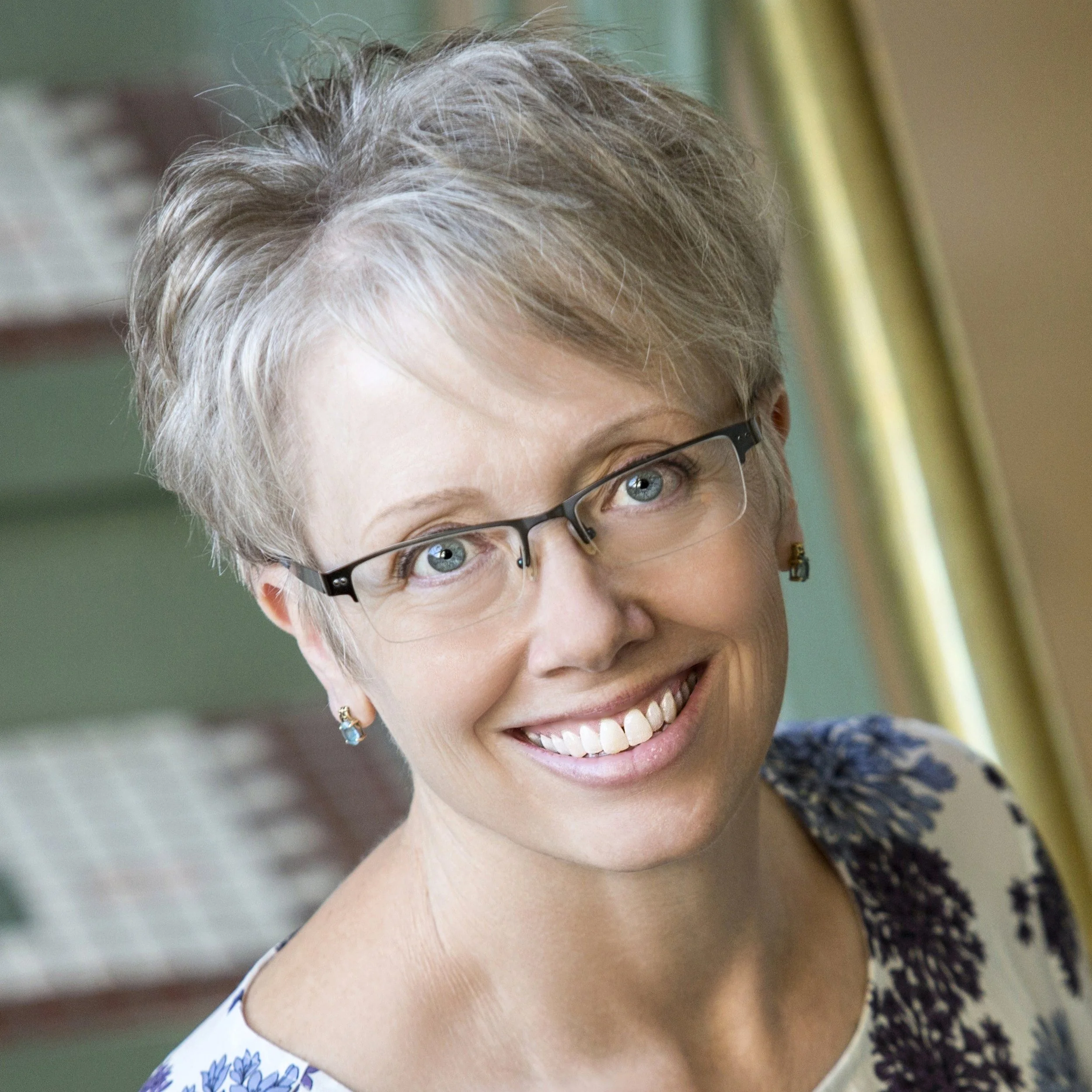 A smiling woman with short gray hair and glasses, wearing earrings and a floral blouse, standing indoors with a green wall and window in the background.