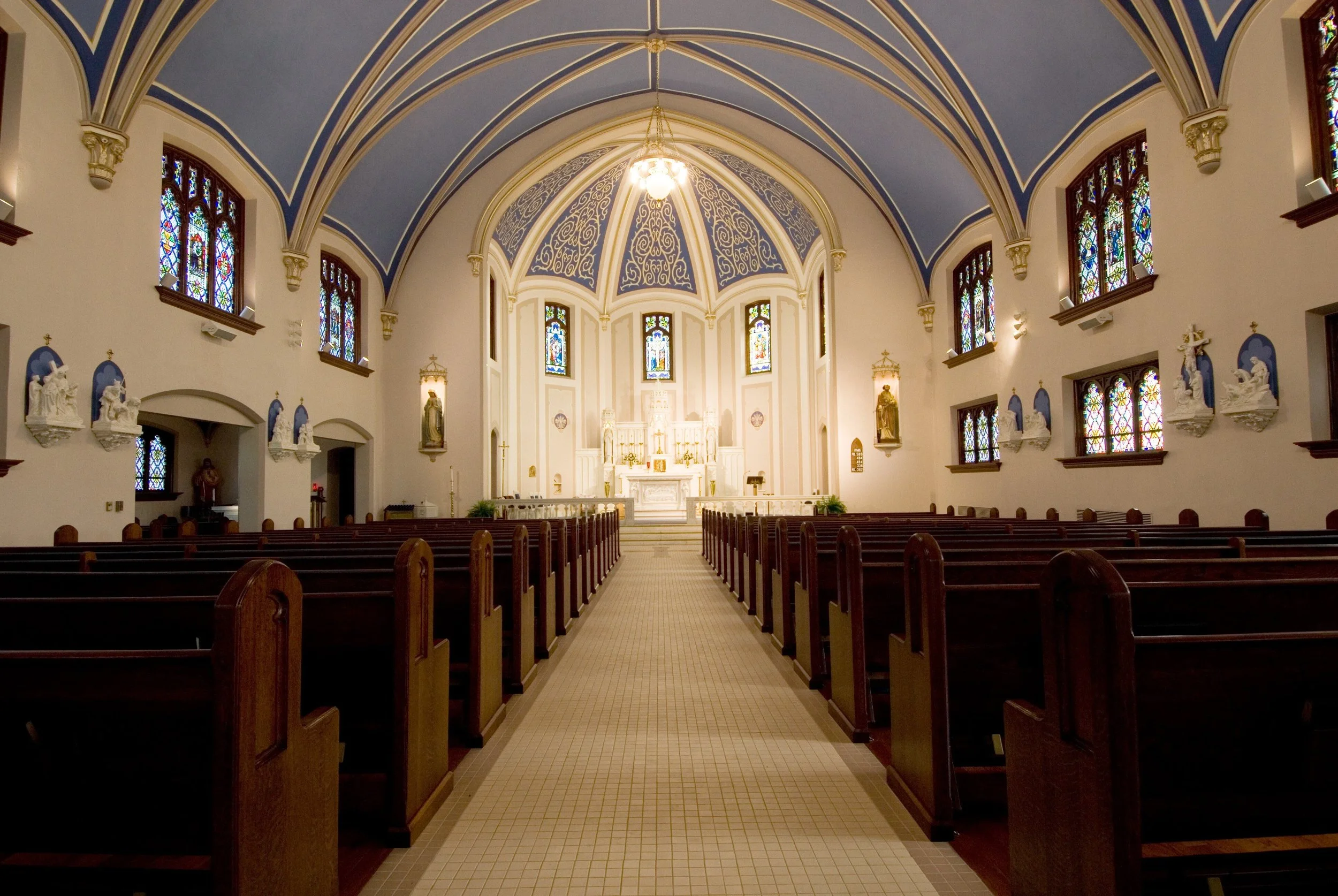 Interior of a church with wooden pews, stained glass windows, a high vaulted ceiling painted blue with gold accents, and an altar at the front.