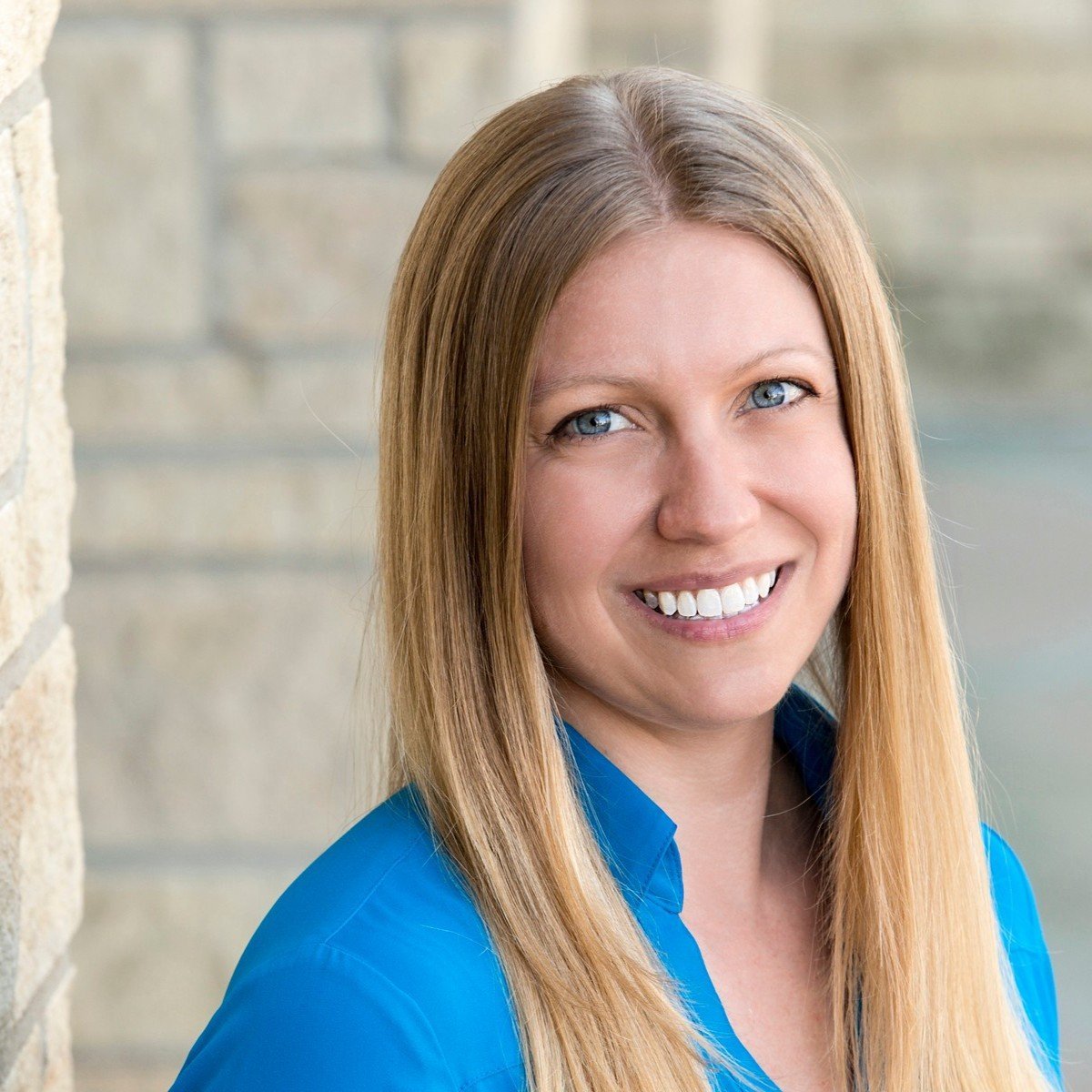 A woman with long blonde hair and blue eyes smiling, wearing a blue shirt, leaning against a brick wall.