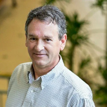 A man with dark hair, wearing a light-colored, checkered button-up shirt, standing indoors with green plants in the background.