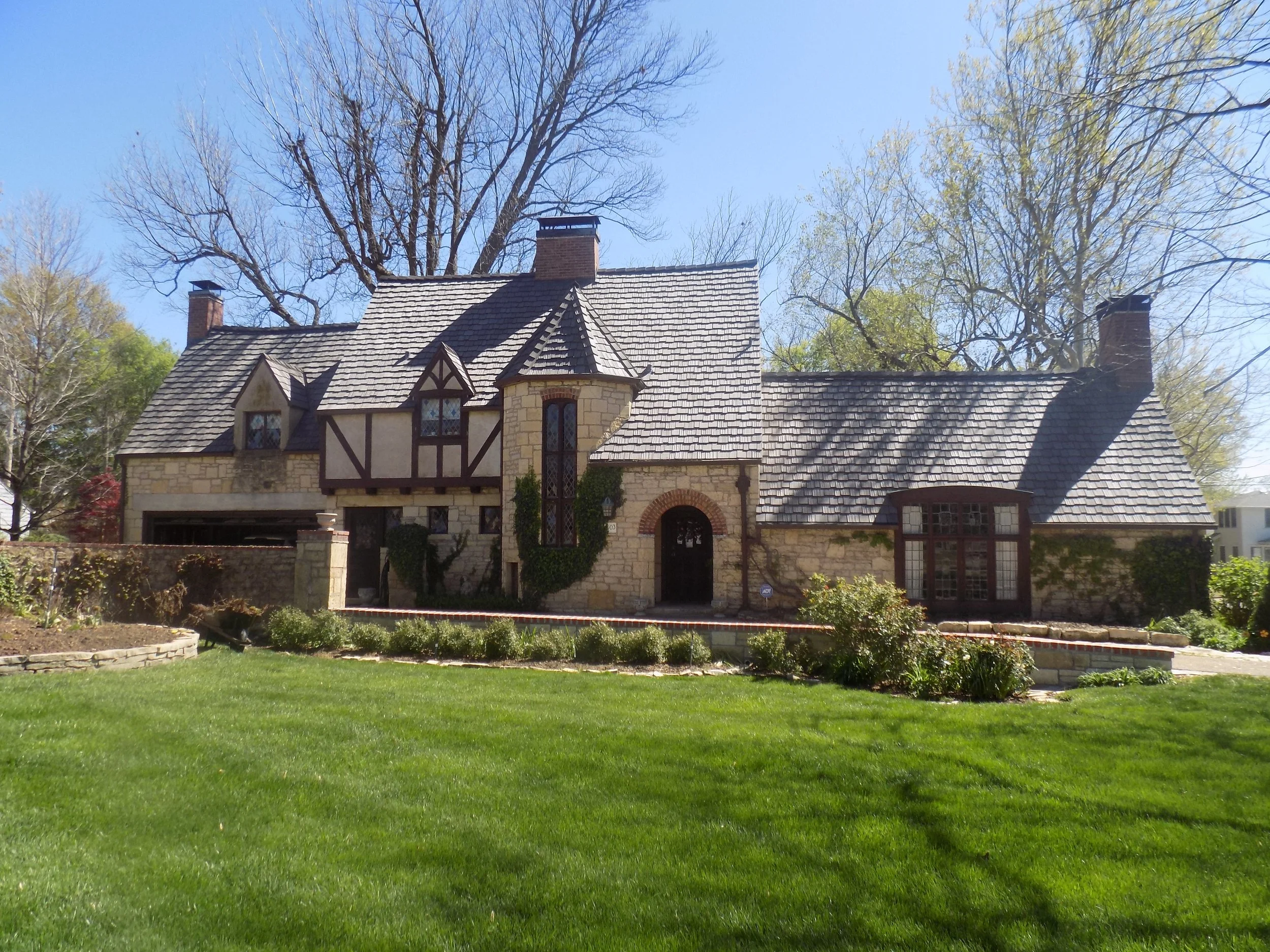 A house with a stone and stucco exterior, steeply pitched roof, multiple chimneys, and a lush green lawn in the foreground, with tall trees and a clear blue sky in the background.