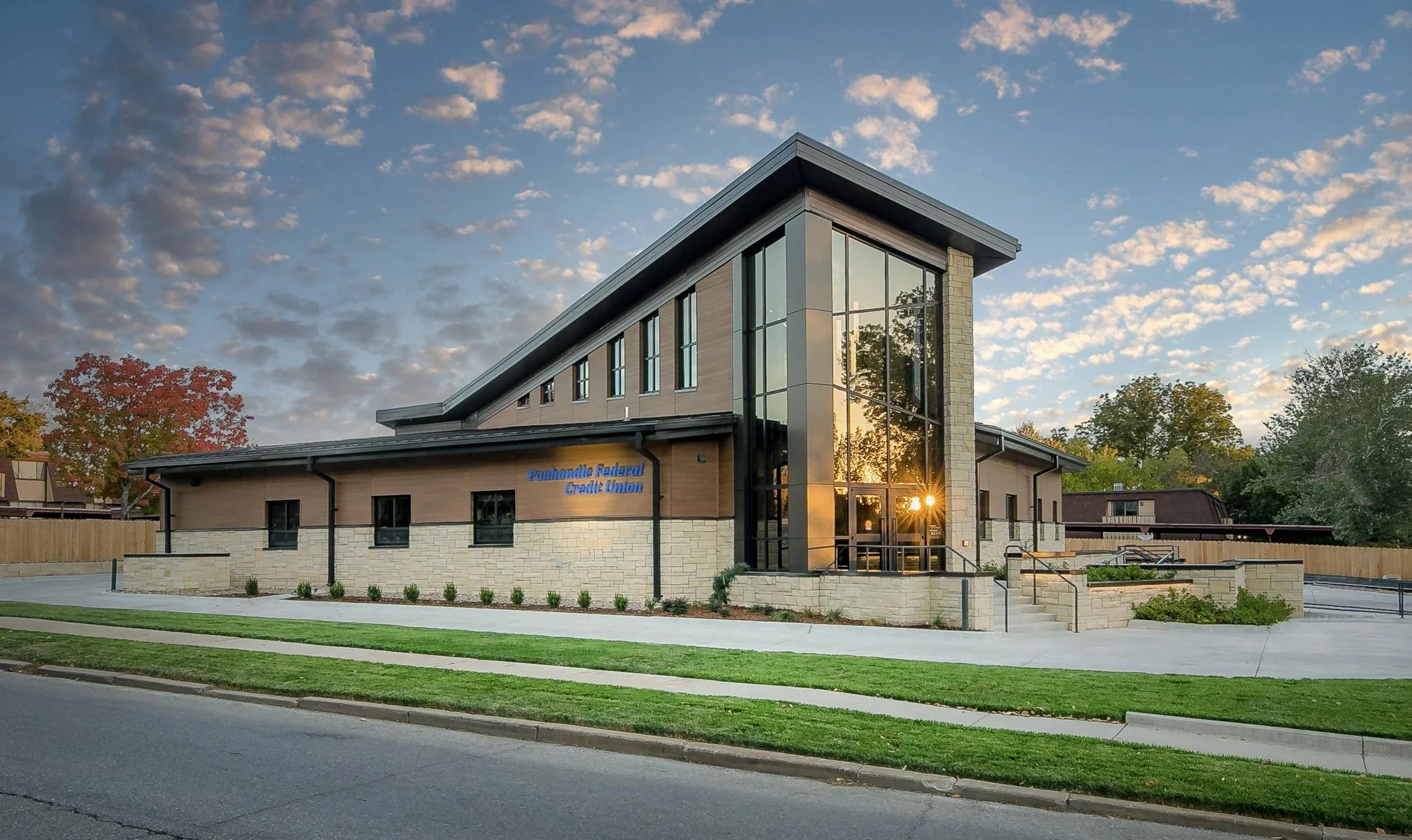 Exterior of the Winfield branch of Panhandle Federal Credit Union with tan brick and large glass windows, located on a street with green grass and sidewalk, during sunset with a partly cloudy sky.