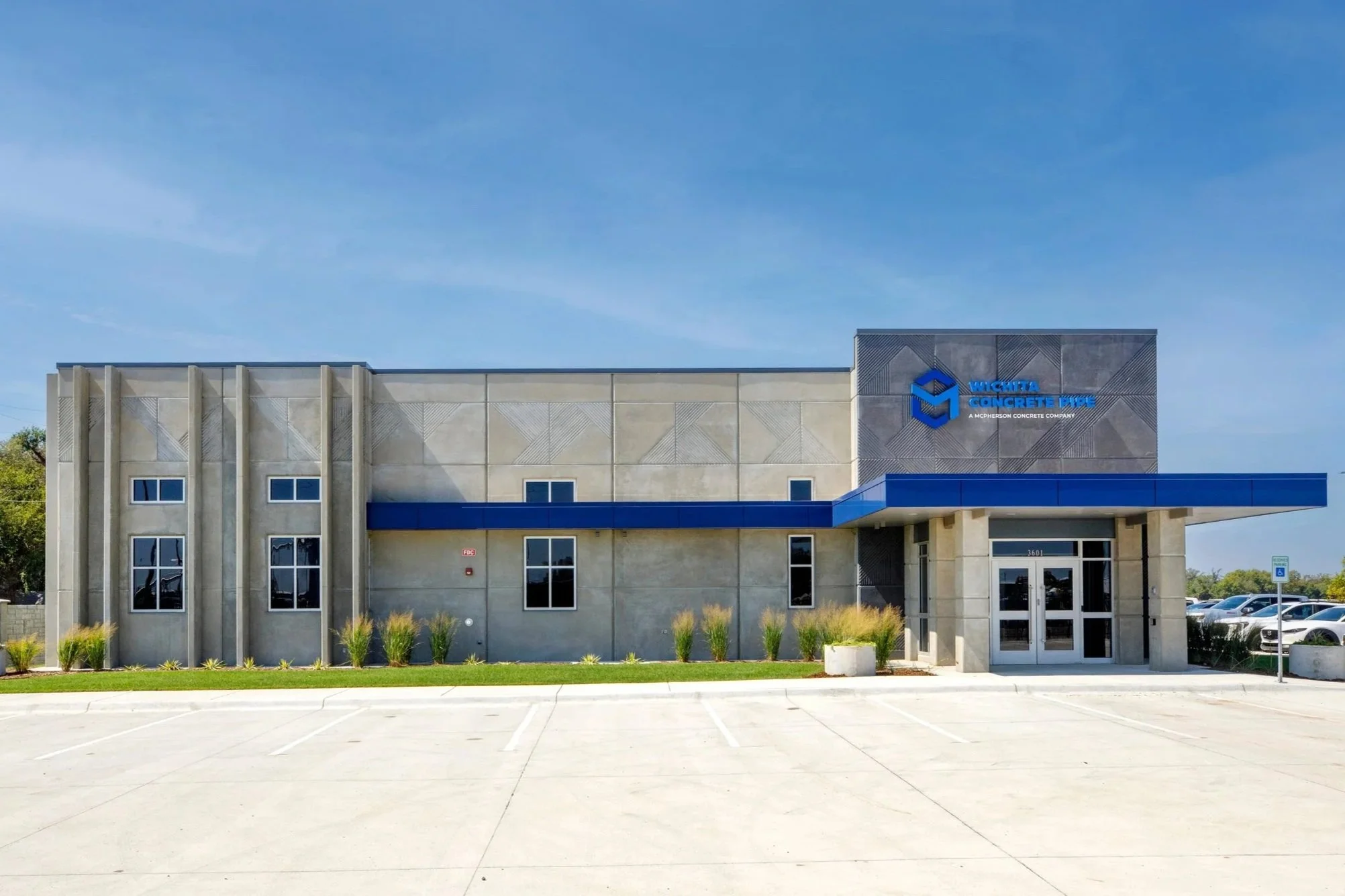 Front view of a commercial building with a sign that reads 'Wichita Concrete Pipe' on the upper right corner. The building has a beige exterior with a blue awning and multiple windows. There are parking spaces in the foreground and some grassy landscaping elements.