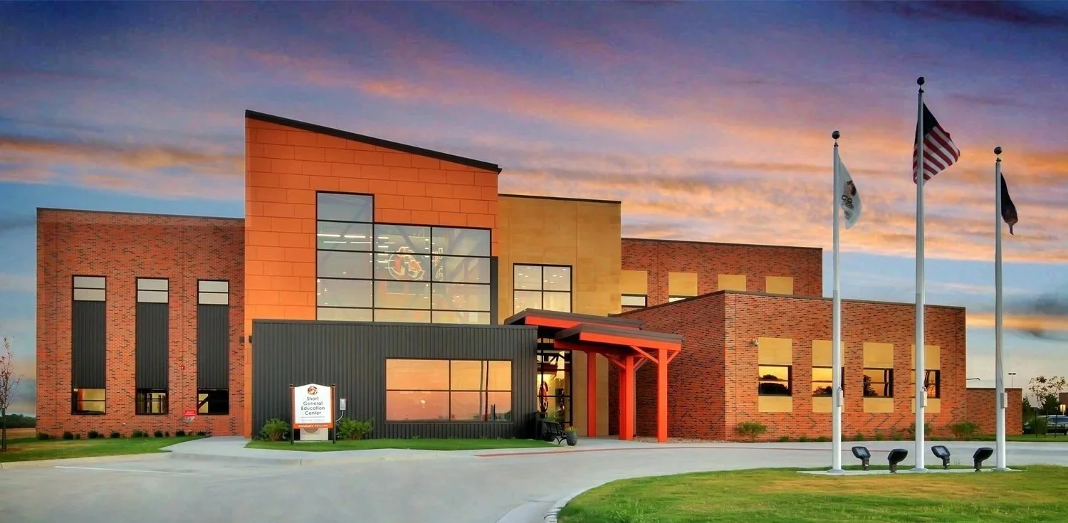 Exterior view of a modern educational center building with brick and metal facade, large glass windows, surrounded by grass, and four flags in front, at sunset.