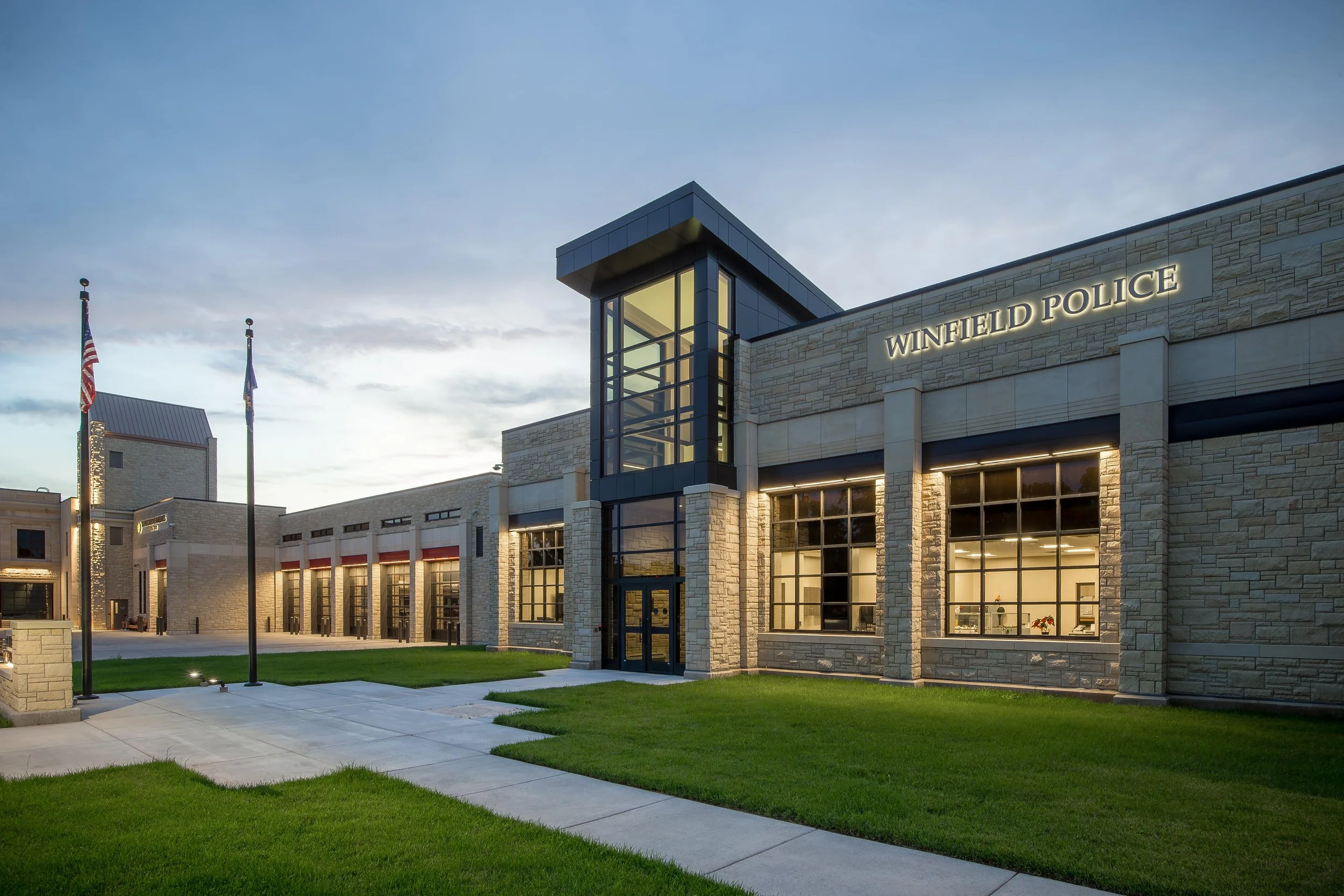 Exterior view of Winfield Police station at dusk, with illuminated sign, large windows, flagpoles with American and state flags, and a manicured lawn.