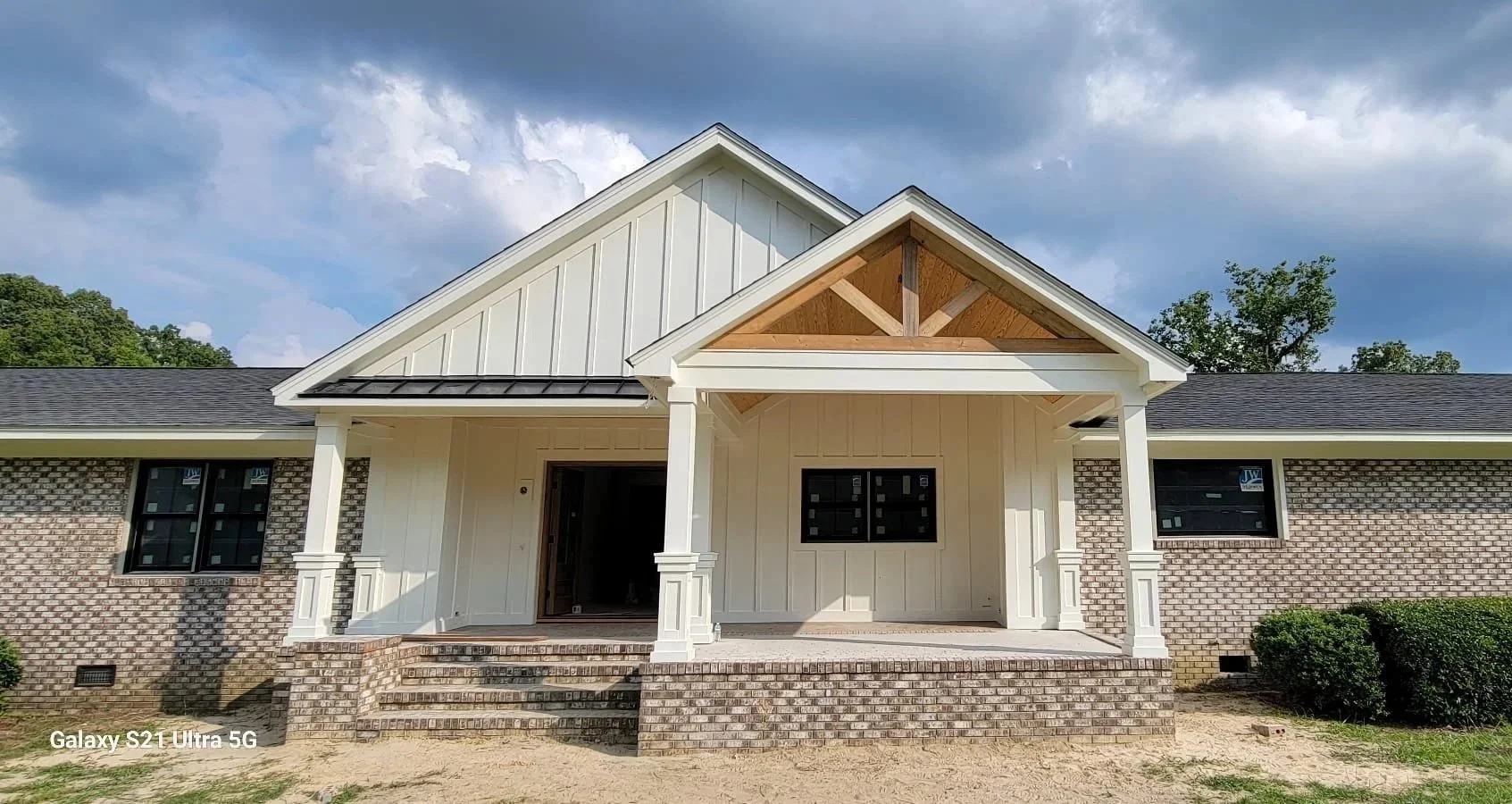 Newly constructed house with white vertical siding, brick foundation, and black window frames, featuring a covered front porch with brick stairs, under a cloudy sky.