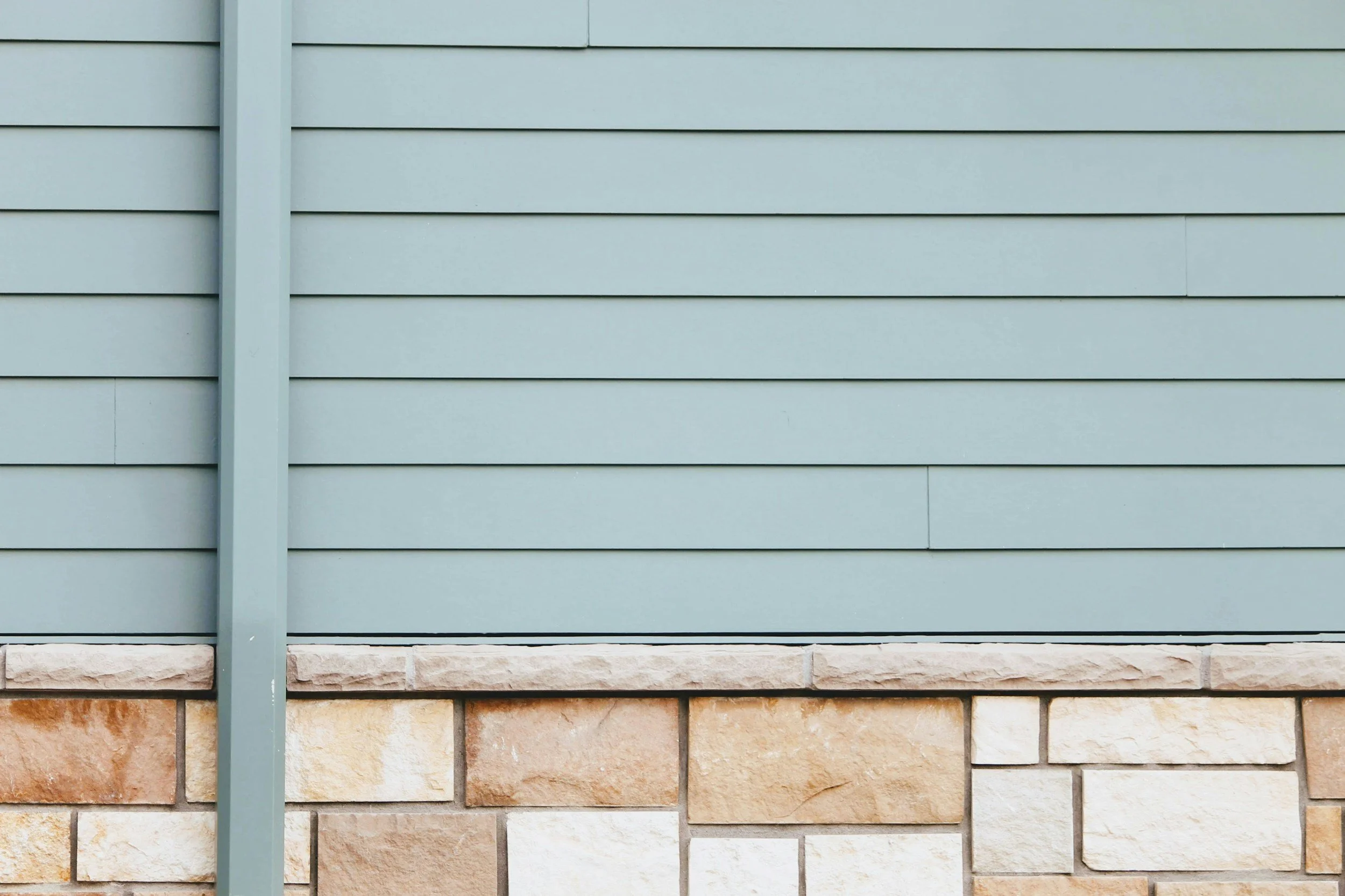 Close-up of a wall with horizontal blue siding on top and a lower section of beige and brown stone bricks, separated by a horizontal line, and a vertical blue corner trim.