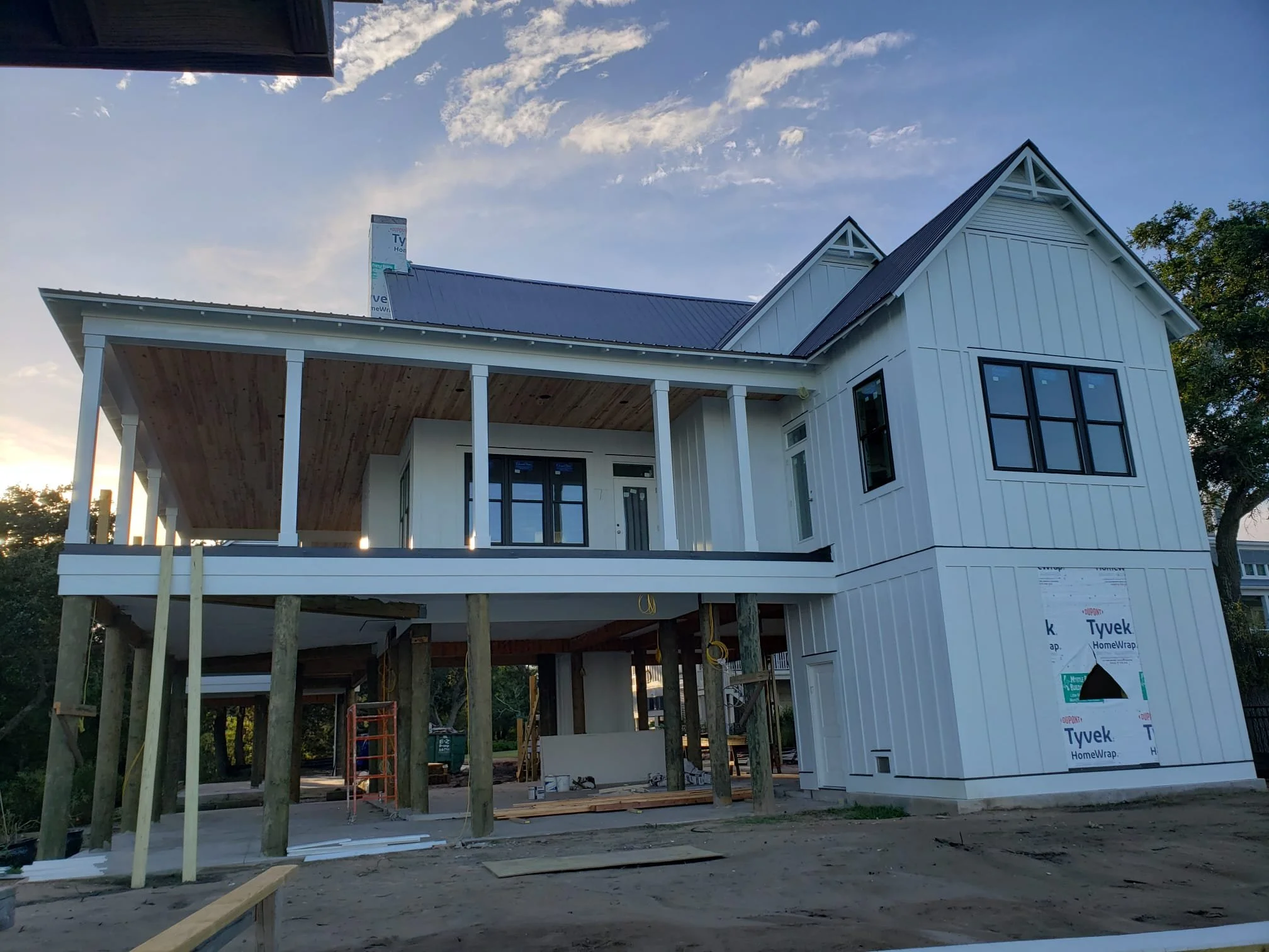 A house under construction with white siding, black windows, and a second-story porch with wood ceiling, supported by wooden beams, at sunset.